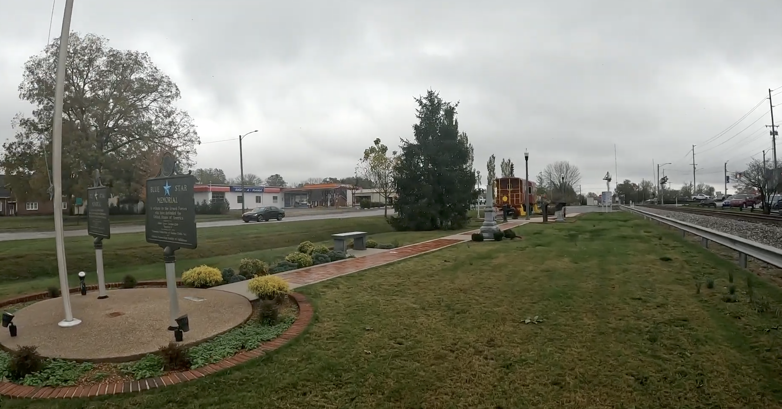 A small park by a railroad track, with a sign memorial, benches, small shrubs, a cement pathway, and a vintage train car on a display track in the background. Overcast sky.