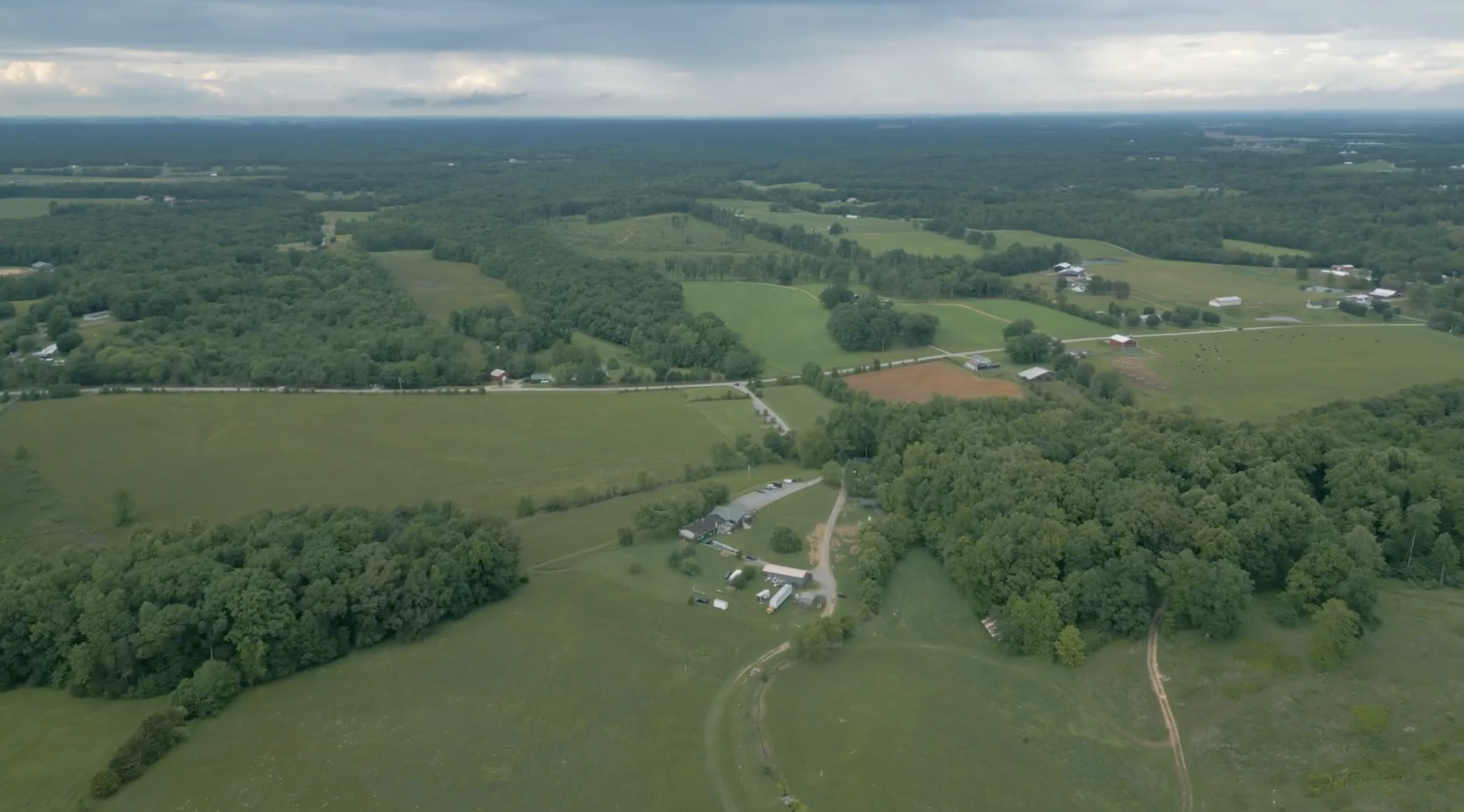 Aerial view of rural farmland with patches of forest, farmhouses, and roads under a cloudy sky.