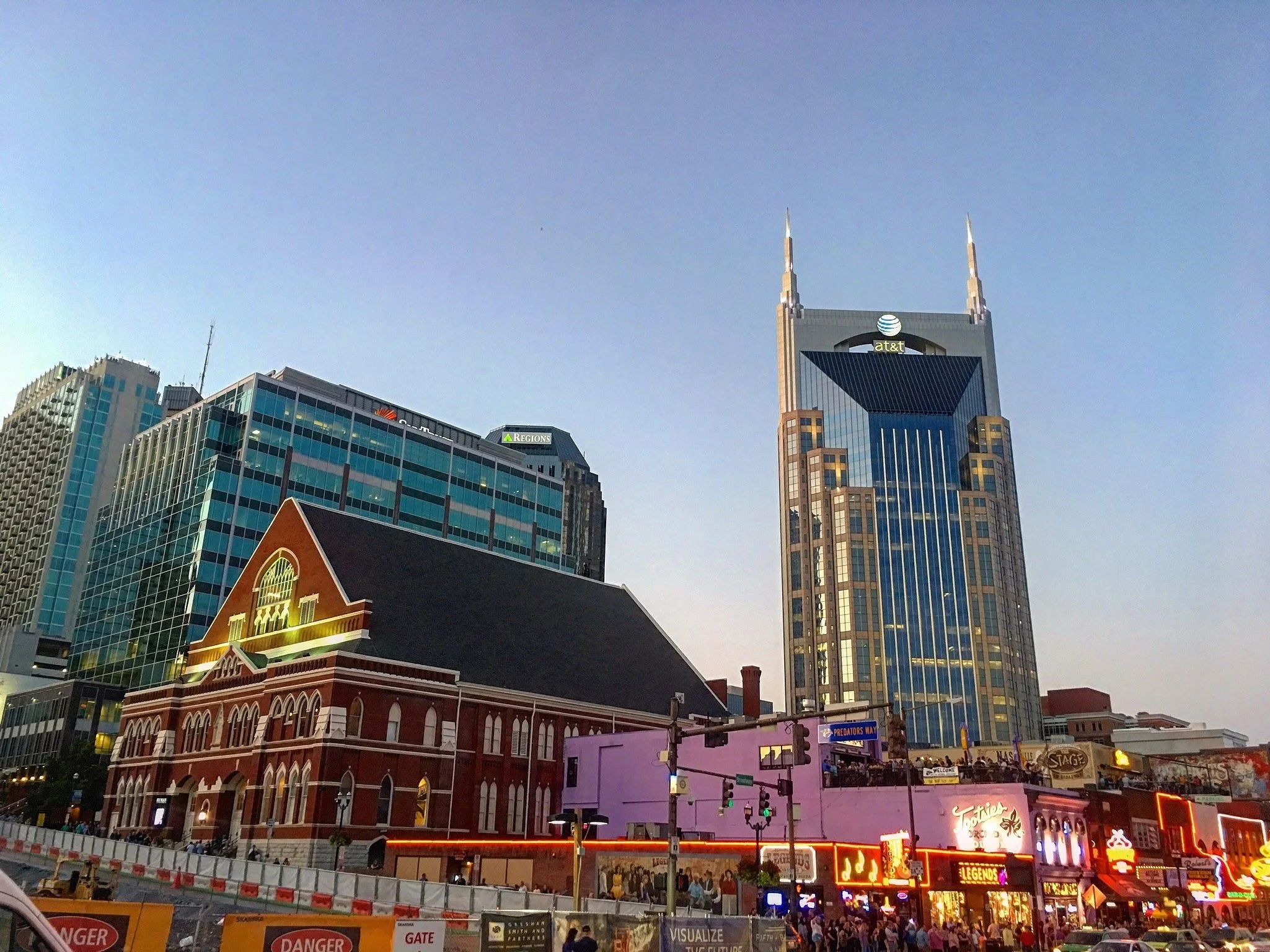 Cityscape with tall modern skyscrapers and a historic red brick building, with neon lights and a crowd of people below