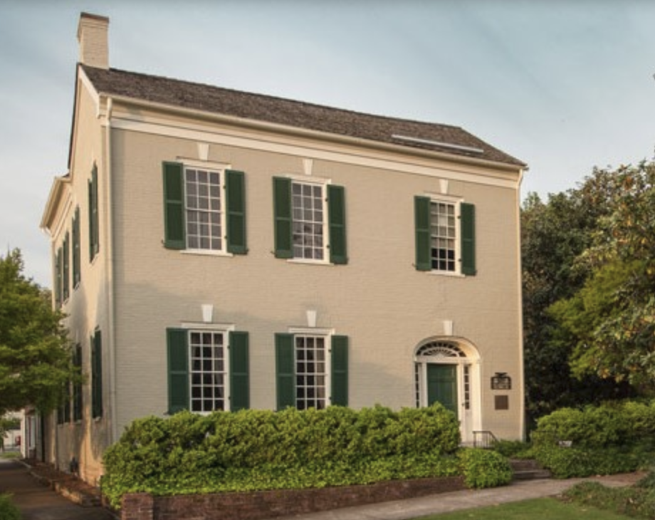 A three-story cream-colored house with green shutters, a white door with a semicircular window above, and surrounded by green bushes and trees.
