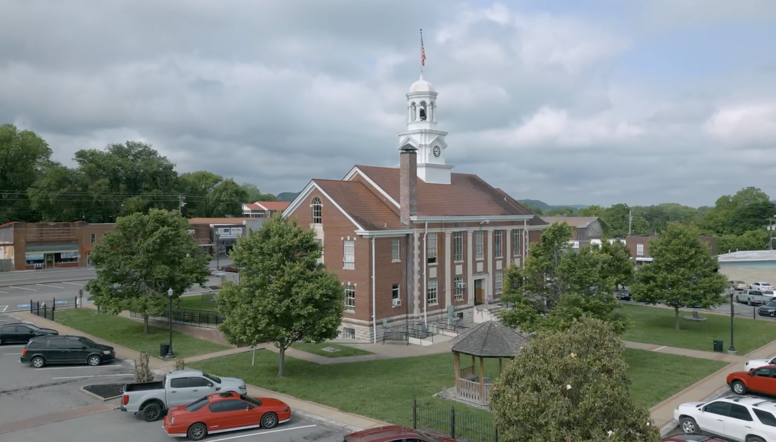 A town square with a historic brick building featuring a white clock tower and a bell, surrounded by parking lots and green trees, with a cloudy sky overhead.