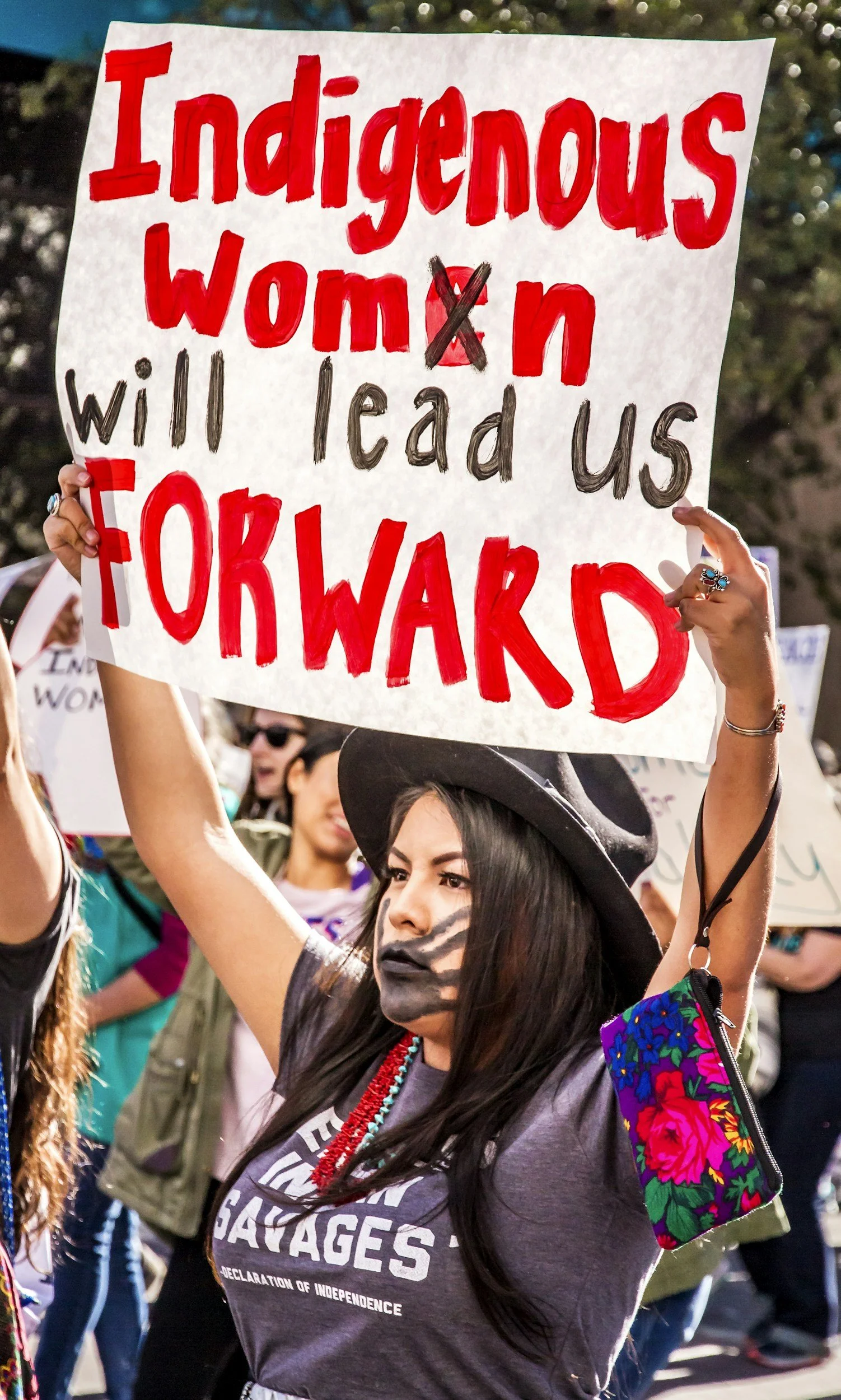 A woman with black face paint holding a sign that reads, 'Indigenous women will lead us forward,' at a protest or rally. She is wearing a dark t-shirt with the word 'sagas' on it and a red beaded necklace, and has a floral-patterned bag on her shoulder. There are other people in the background.