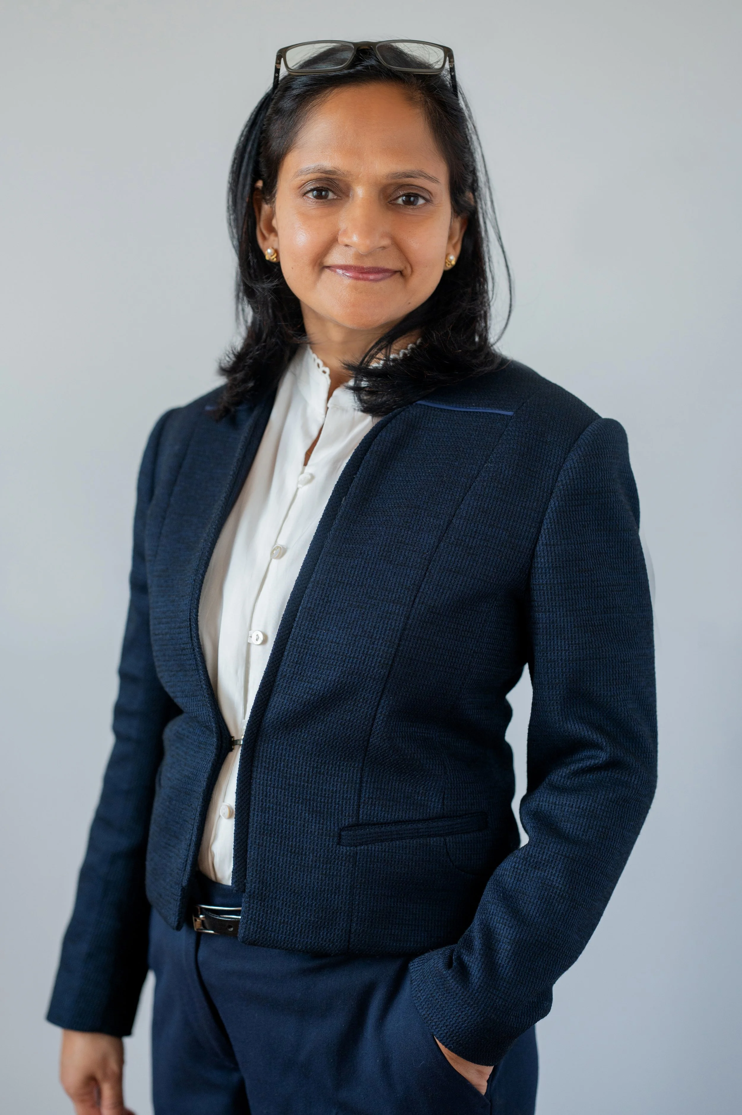 Professional woman with glasses on her head, dressed in a navy blazer over a white shirt, posing against a plain light gray background.