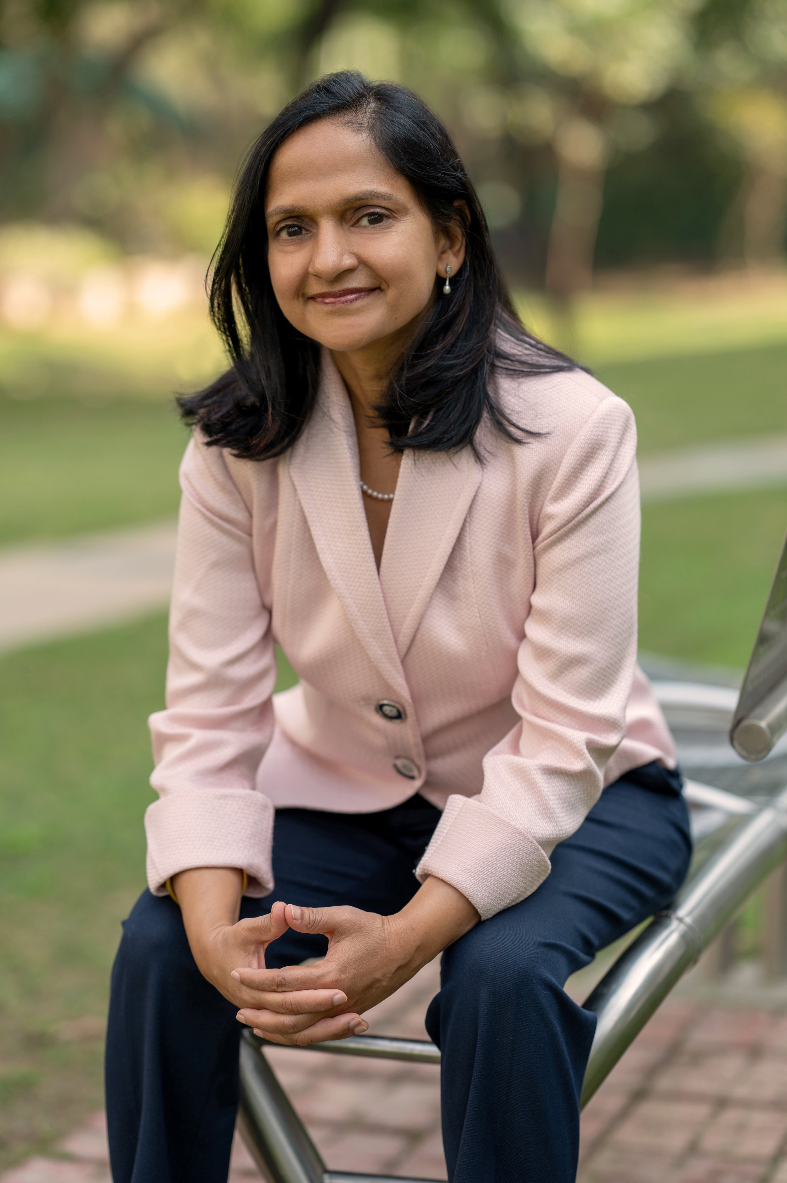 A woman with black hair and earrings sitting on a metal bench outdoors, wearing a light pink blazer and dark pants, smiling with a blurred green park background.