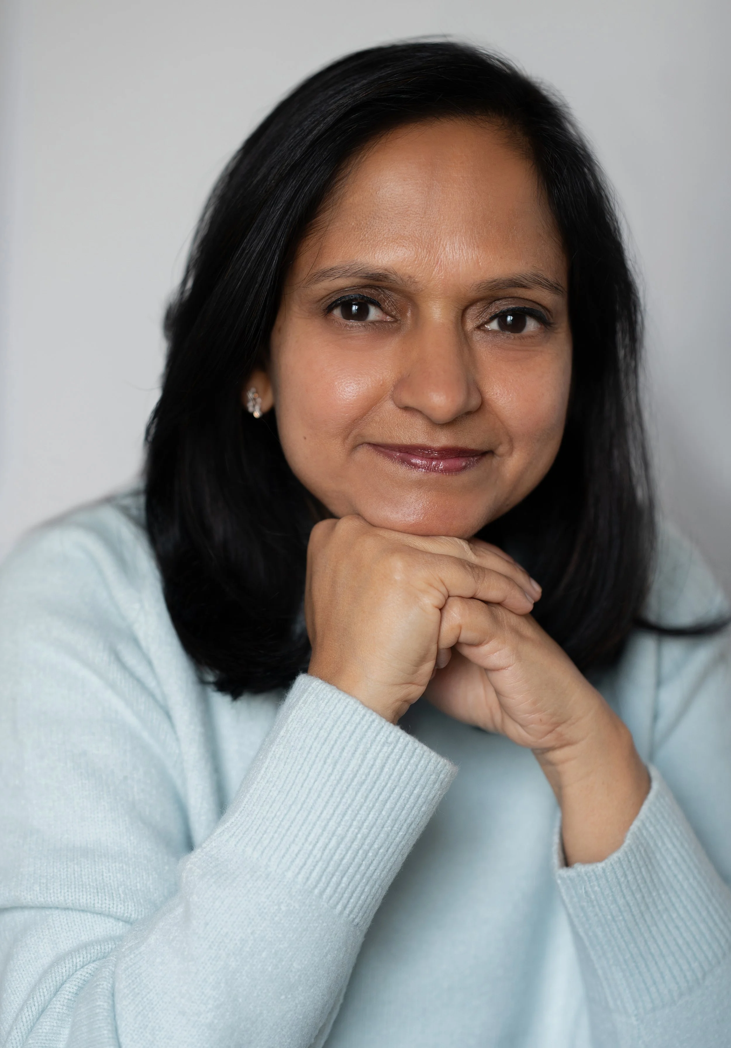 Close-up of a woman with black hair, wearing a light blue sweater, smiling softly with her chin resting on her hands, in front of a plain gray background.