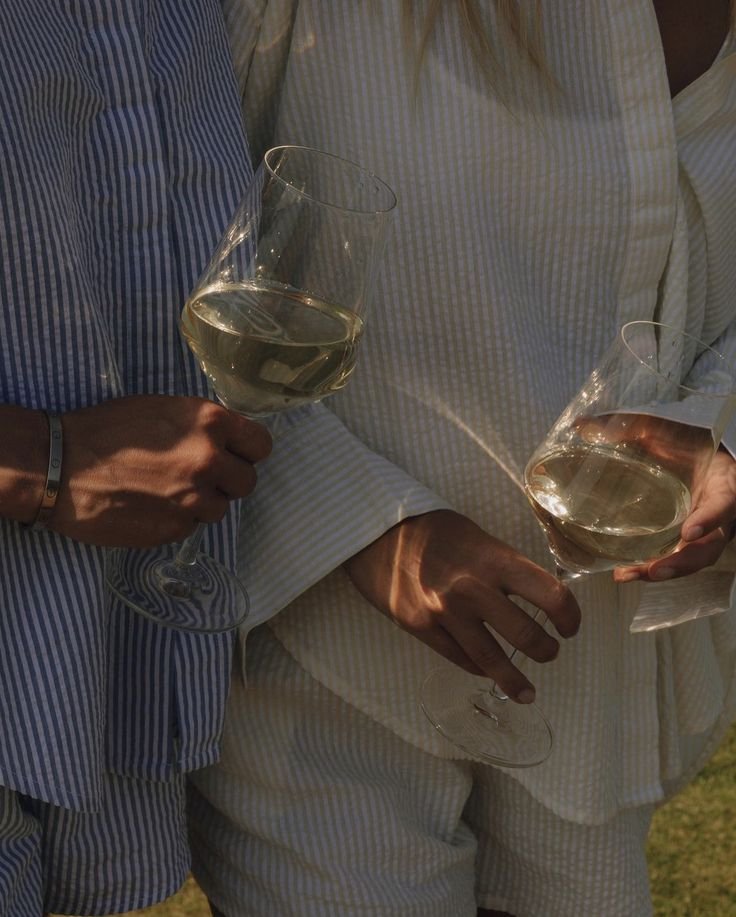Two people holding wine glasses filled with white wine, dressed in light-colored clothing, standing close together outdoors.