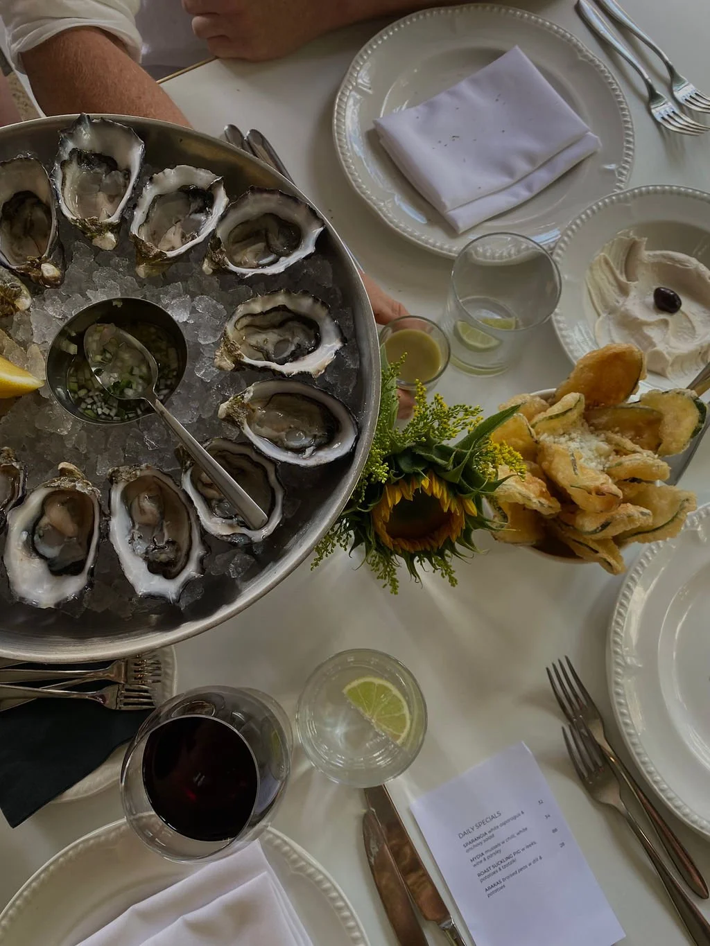 Table set with oysters on ice, lemon wedges, a glass of red wine, a cocktail with a lemon slice, fried artichoke hearts, a plate with a black olive and creamy dip, a sunflower in a small bouquet, and neatly folded white napkins.
