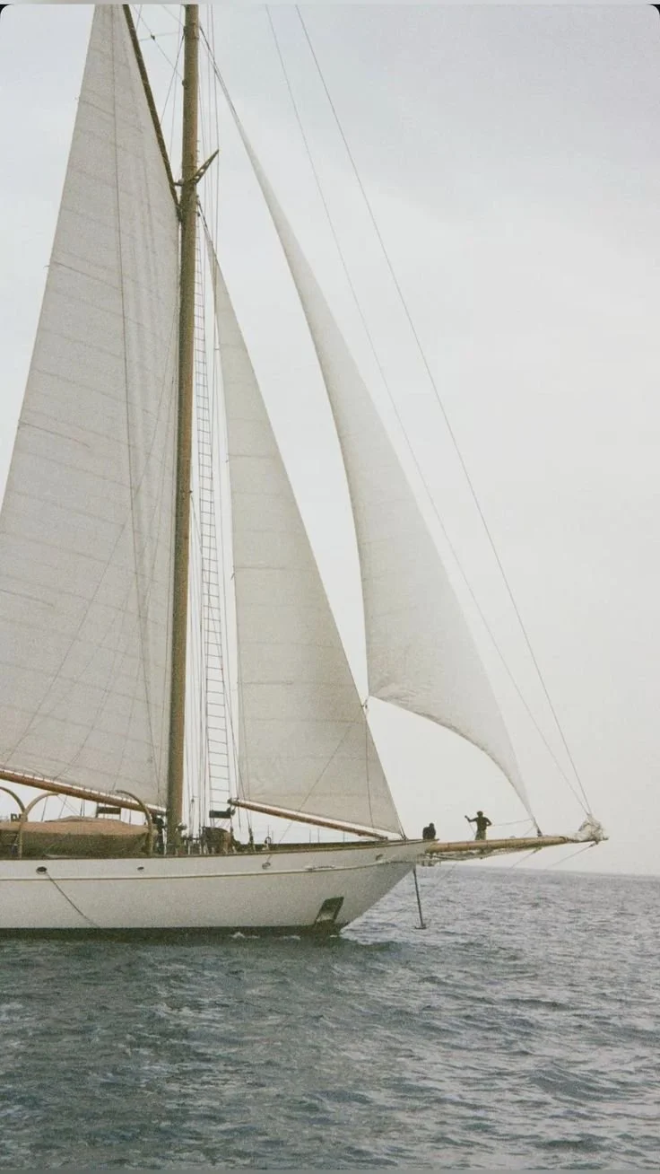 A large sailing yacht with multiple white sails on open water, two people standing on the bow, and the sky above appears cloudy.