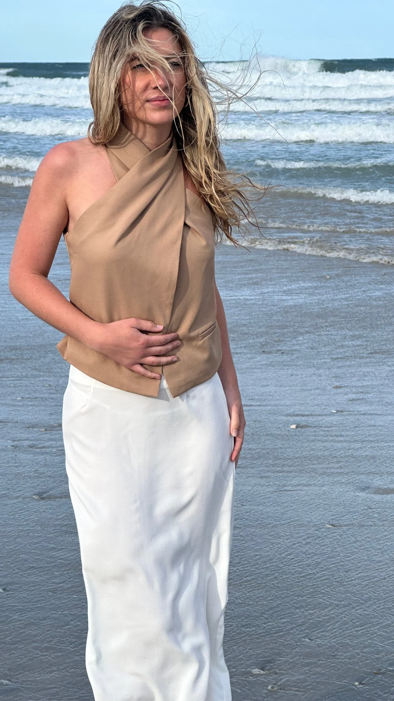 Woman standing on the beach with ocean waves in the background, wearing a beige halter top and a white skirt, with wind blowing her blonde hair.