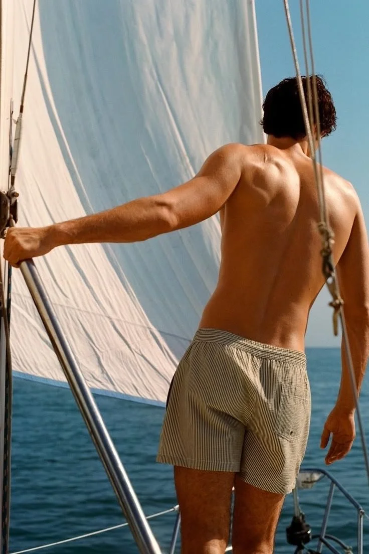 A shirtless man with dark curly hair steering a sailboat on the ocean, wearing beige striped shorts.