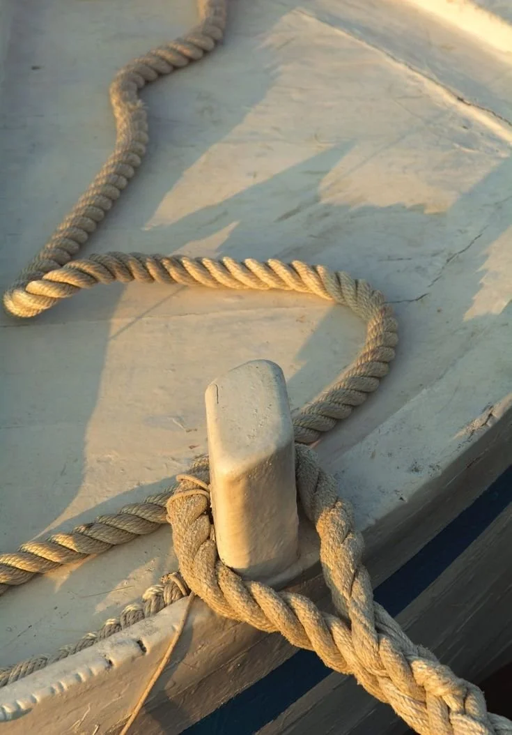 Close-up of a heavy-duty rope tied around a white bollard on a boat, with sunlight casting shadows on the boat's surface.