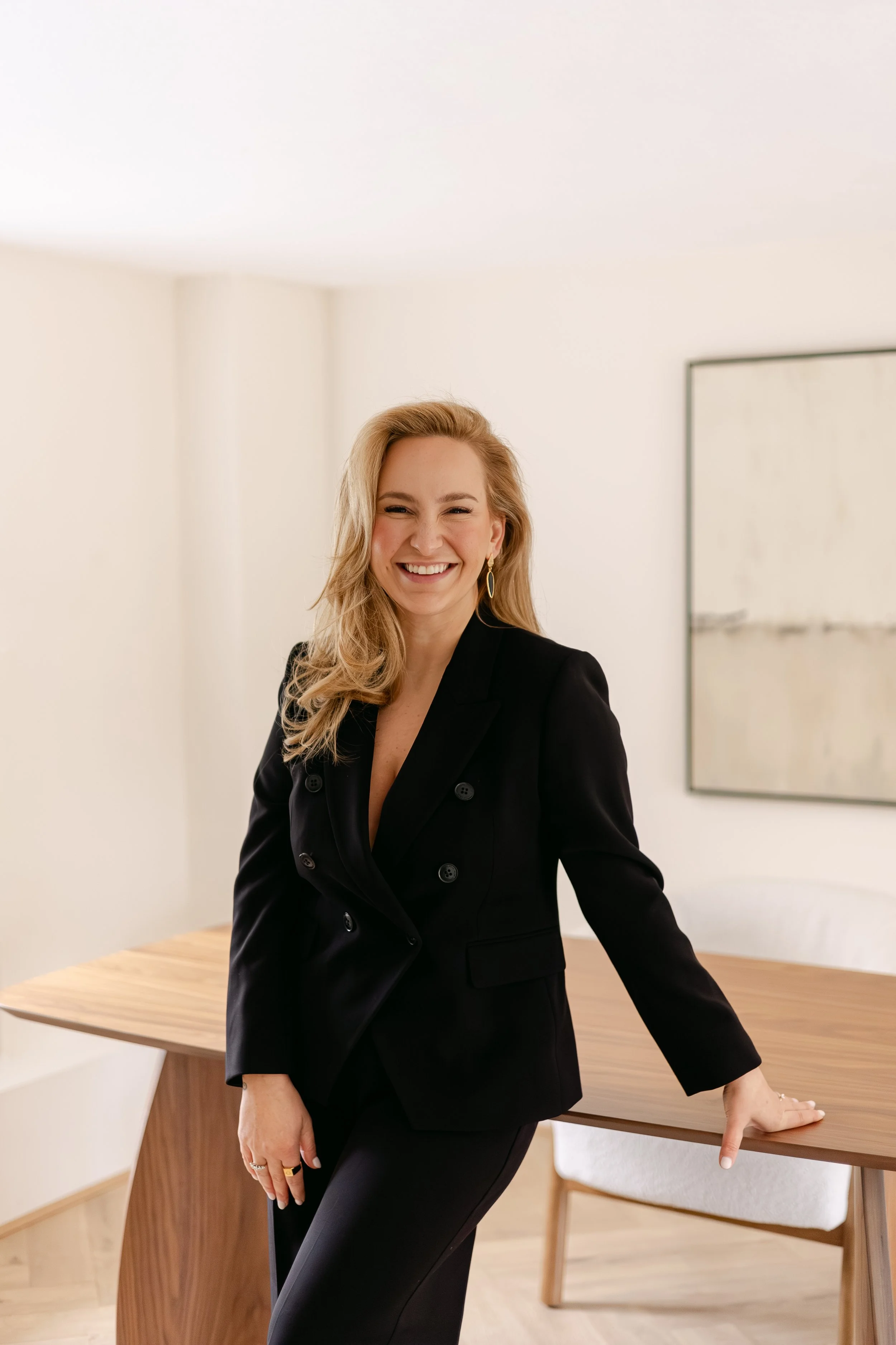 A woman with blonde hair wearing a black blazer and pants, smiling and leaning on a wooden table in a minimalistic room with white walls and abstract artwork.
