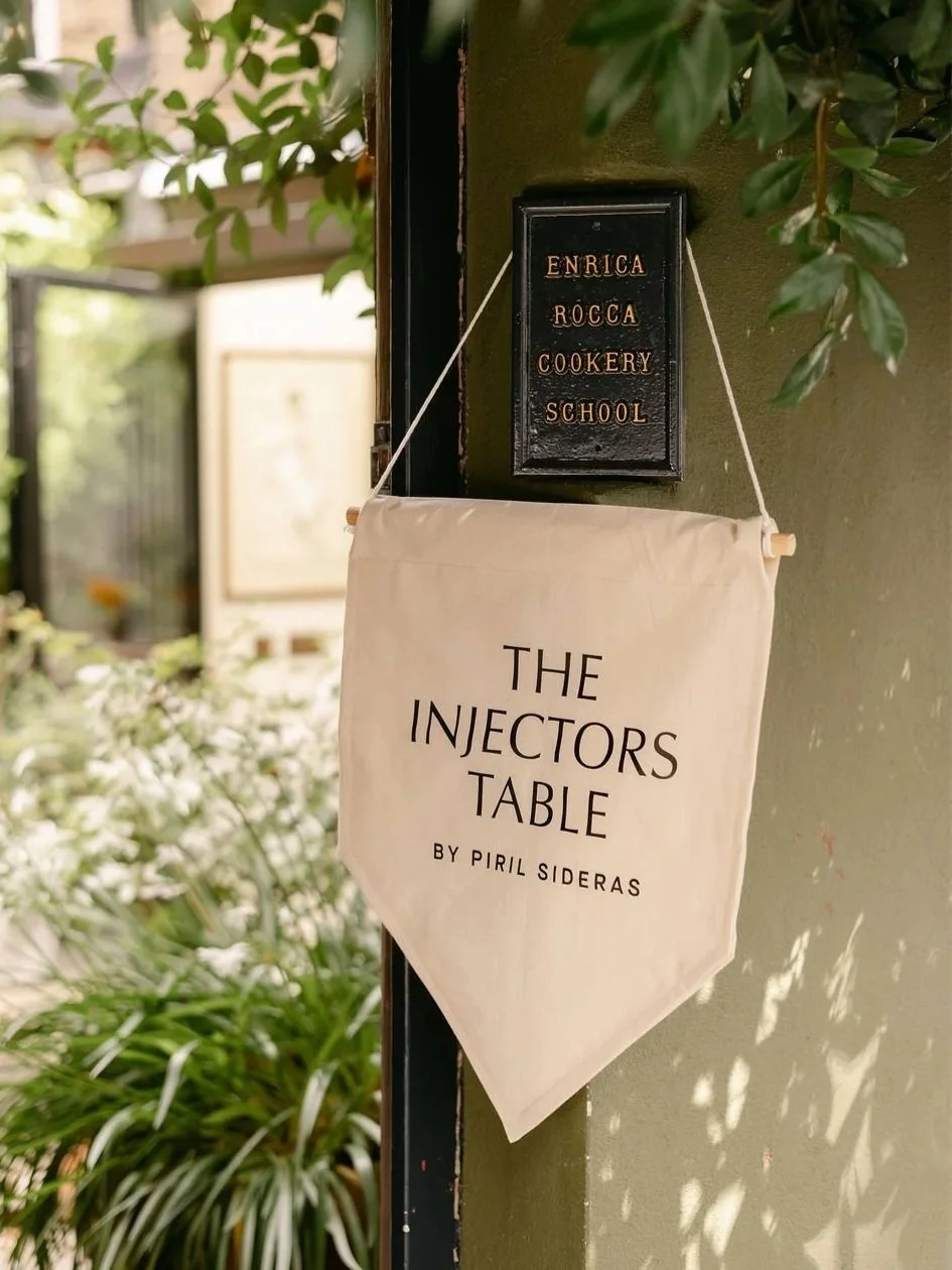 A black plaque with copper-colored text reads 'Enrica Rocca Cookery School,' attached to a green wall. Below, a beige fabric banner with black text says 'The Injectors Table by Piril Sideras,' hanging from a wooden dowel and string. In the background, plants and a window are visible.