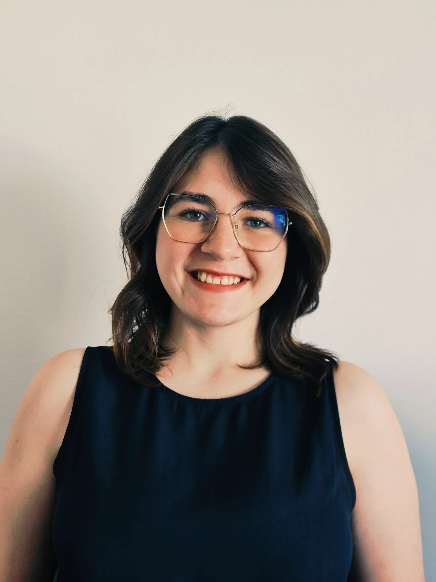 Mulher com cabelos castanhos escuros, usando óculos e um vestido preto, sorrindo diante de uma parede bege.