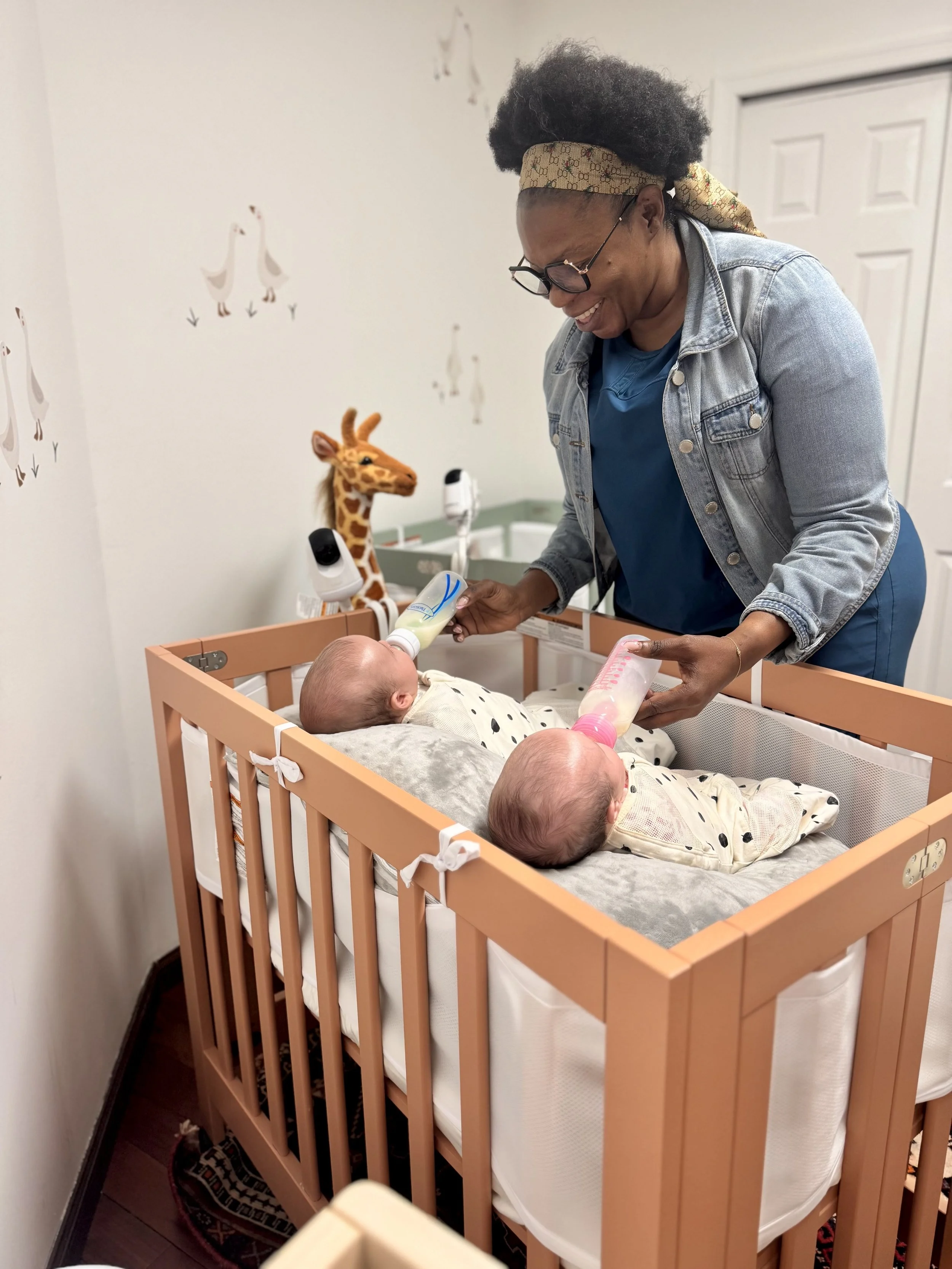 A woman with glasses, curly hair, and a patterned headscarf feeding a baby with a bottle in a crib with gender-neutral bedding and a giraffe plush toy nearby, in a nursery with wall decals of geese.