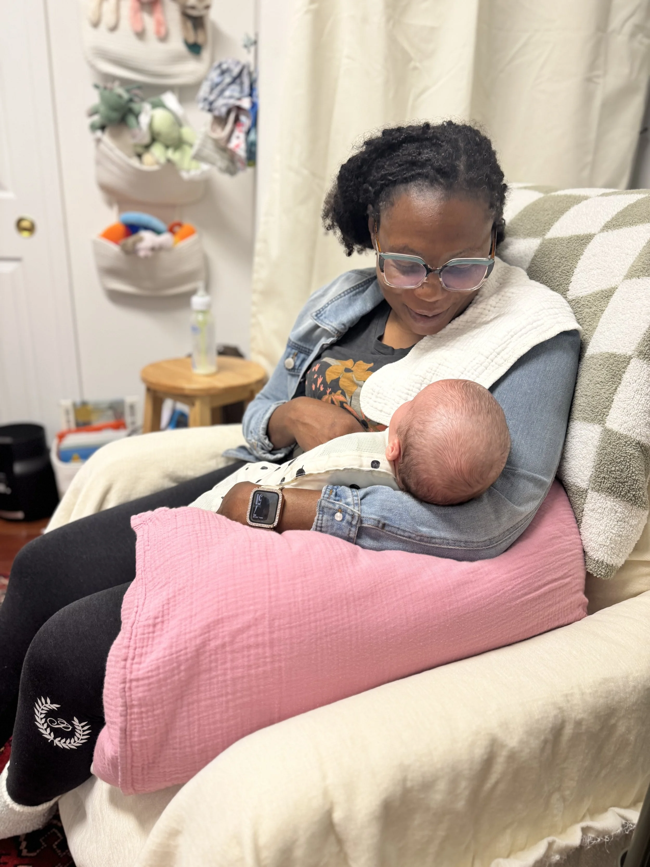 A woman with short curly hair and glasses holding a newborn baby in a cozy room. The woman is sitting on a couch with a pink cushion across her lap. The room has a small wooden side table with a bottle and other items, a closet with hanging clothes and plush toys, and cream-colored curtains.