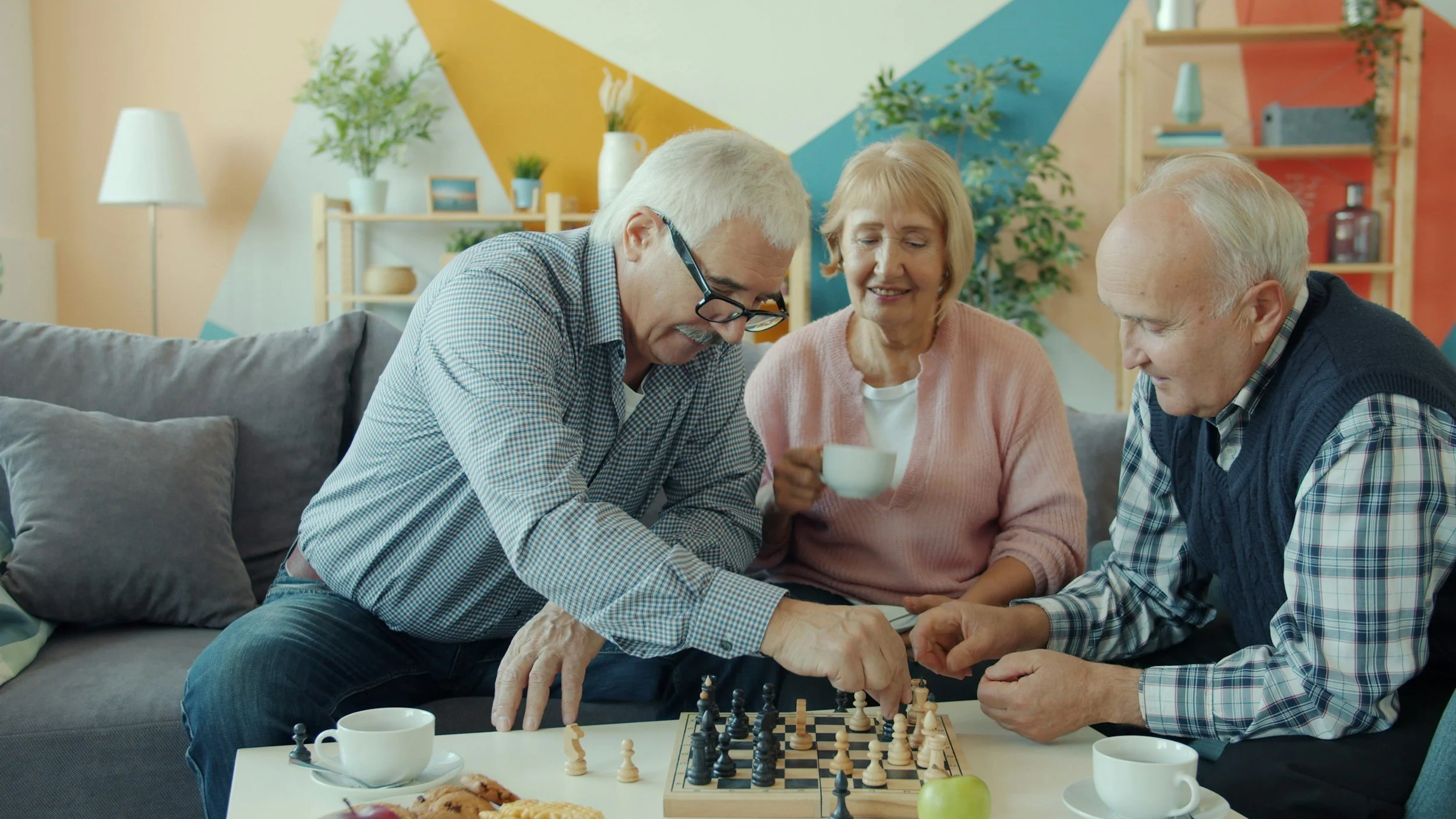 Four elderly people are playing chess together on a white table in a cozy, colorful living room. There are cups of coffee and some food on the table.