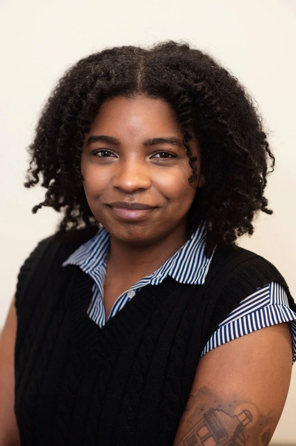 Close-up of a woman with dark curly hair, wearing a striped shirt and a black sweater vest, smiling softly with a tattoo visible on her upper arm, against a plain light background.