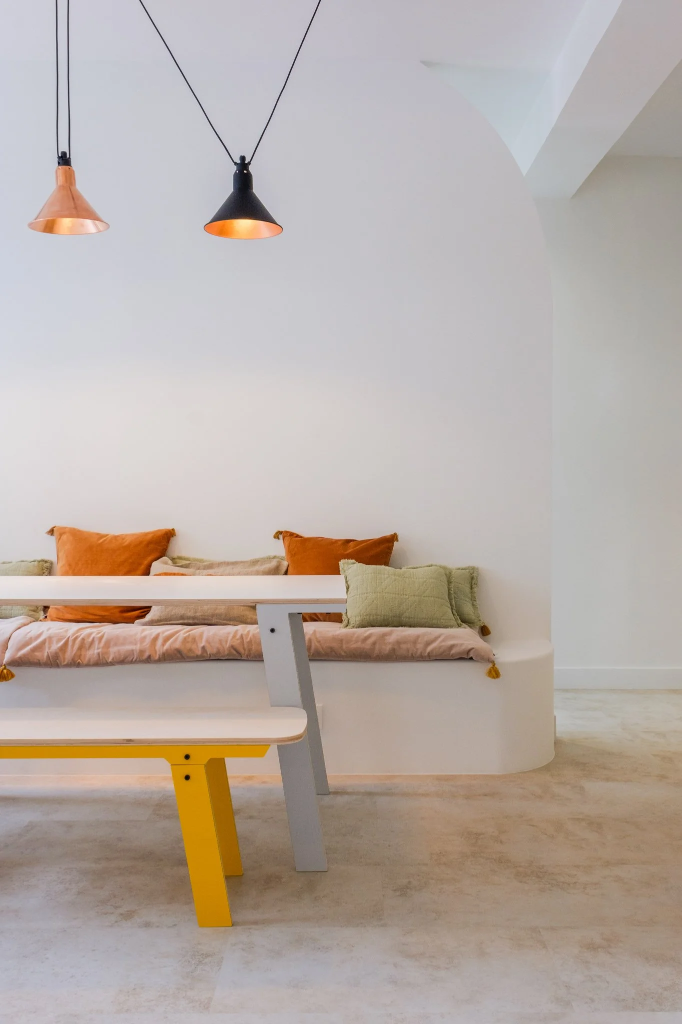 Un salon minimaliste avec un canapé beige clair, décoré de coussins orange et vert, sous un plafond blanc avec deux lampes suspendues, une noire et une cuivre.