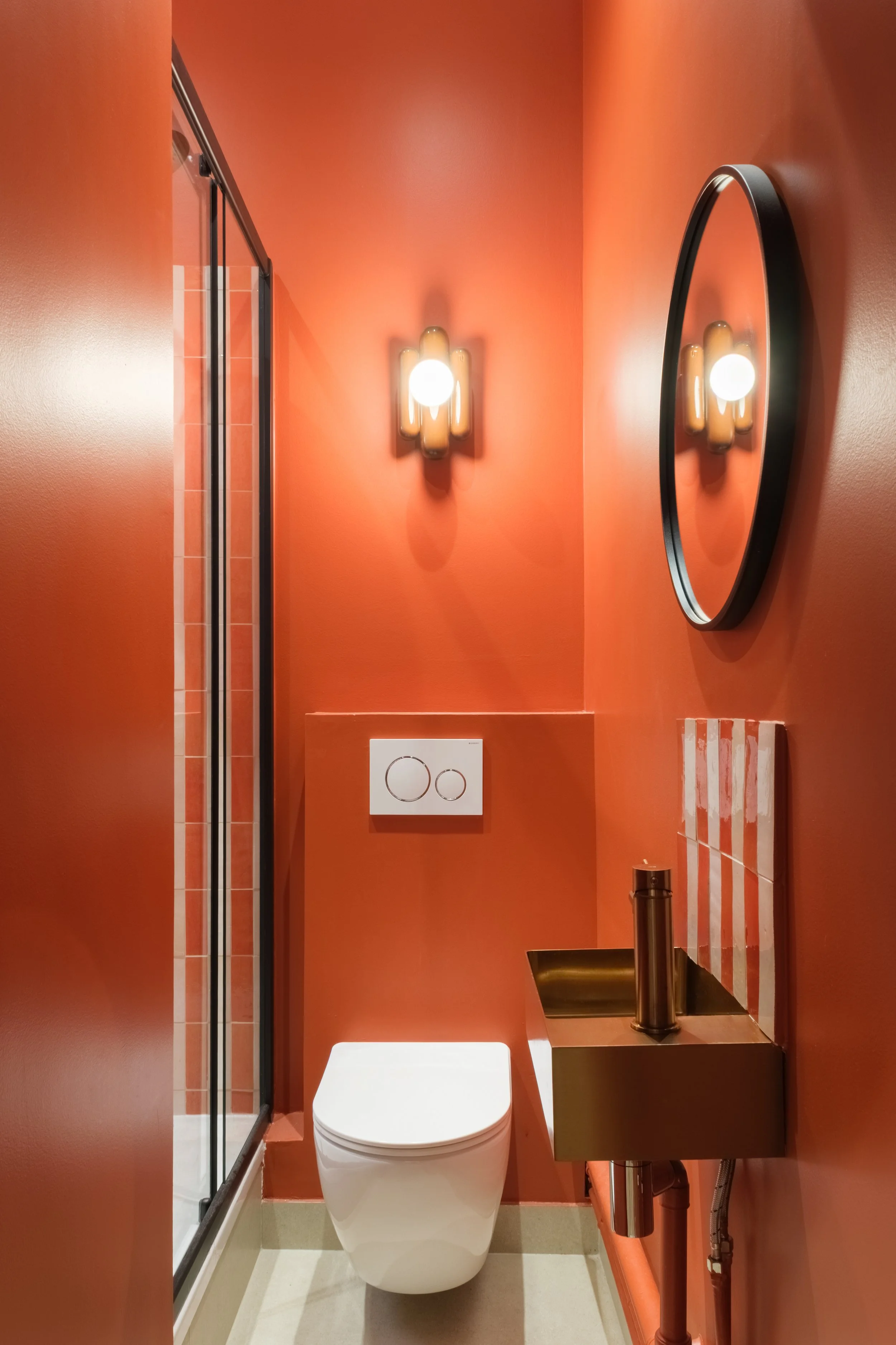 Salle de bain avec murs orange, toilette moderne, miroir rond, lavabo en métal, lumière murale, porte de douche en verre, accessoires de salle de bain.