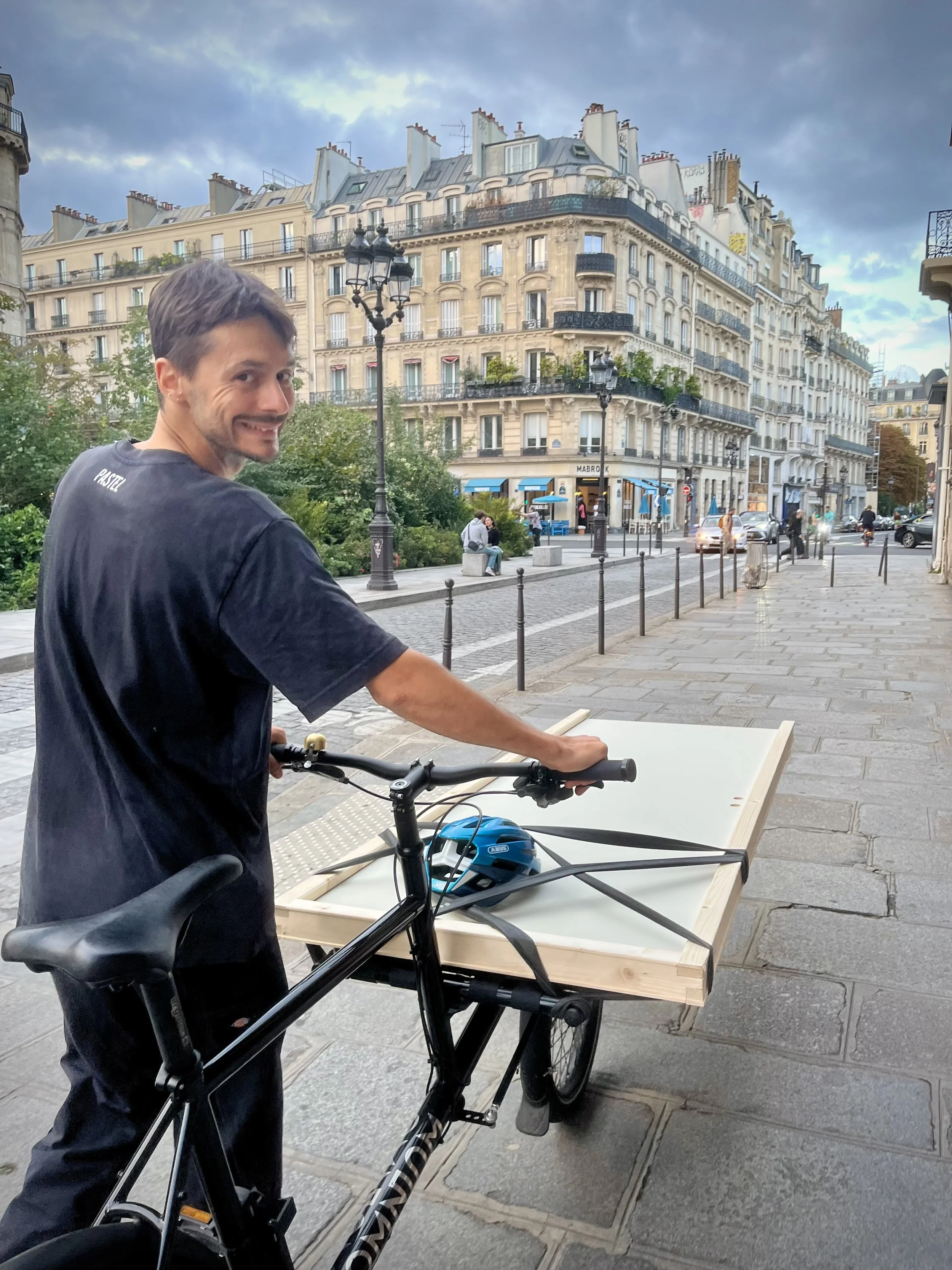Un homme souriant avec un vélo, se tenant à côté d'une table en bois sur une rue parisienne avec de beaux bâtiments haussmanniens en arrière-plan, par temps légèrement nuageux.