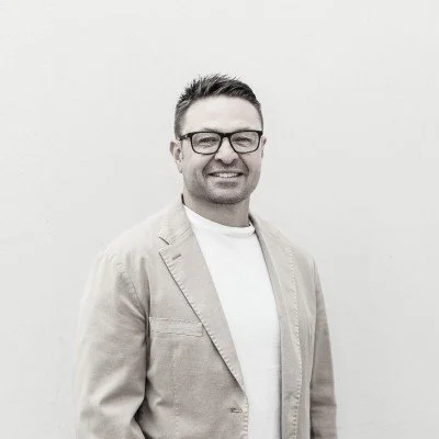 A man with glasses and short dark hair, smiling, wearing a beige blazer over a white shirt, standing against a plain white background.