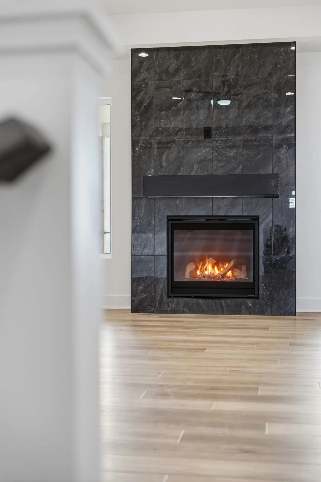 A modern living room with a black marble fireplace, a small window on the white walls, and light wood flooring.