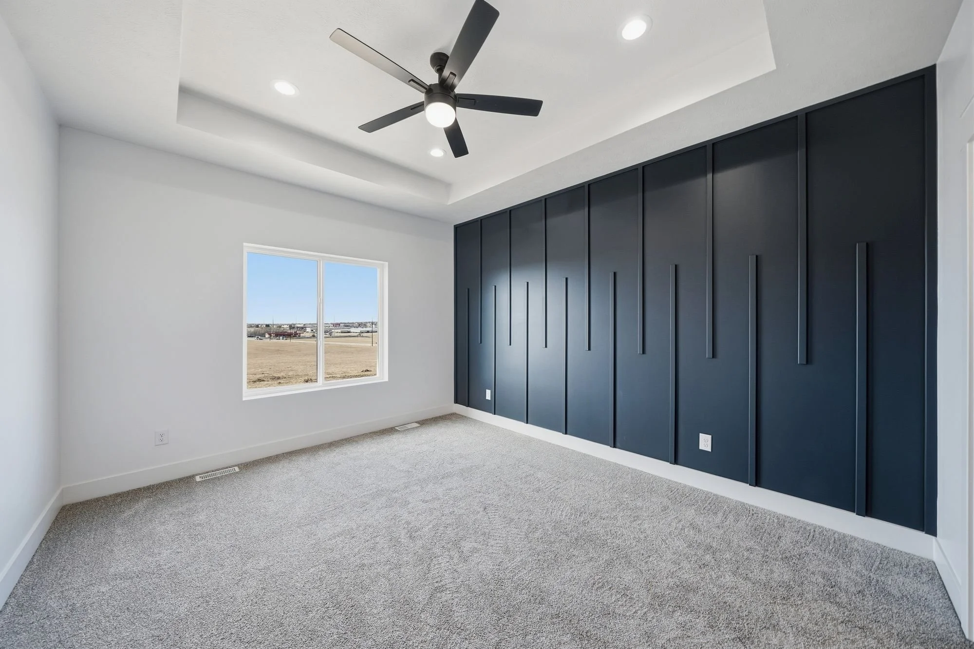 Empty room with white walls, beige carpet, a large window showing an outdoor view of a field and sky, a black ceiling fan, and black storage cabinets along one wall.