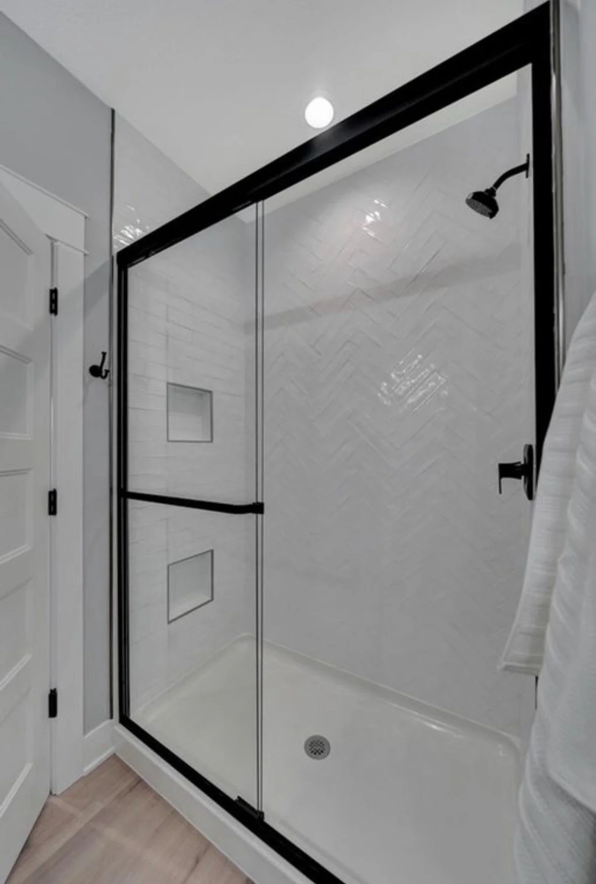 A modern bathroom shower with clear glass doors, black hardware, white tiled walls in a herringbone pattern, a built-in niche, and a ceiling-mounted showerhead.