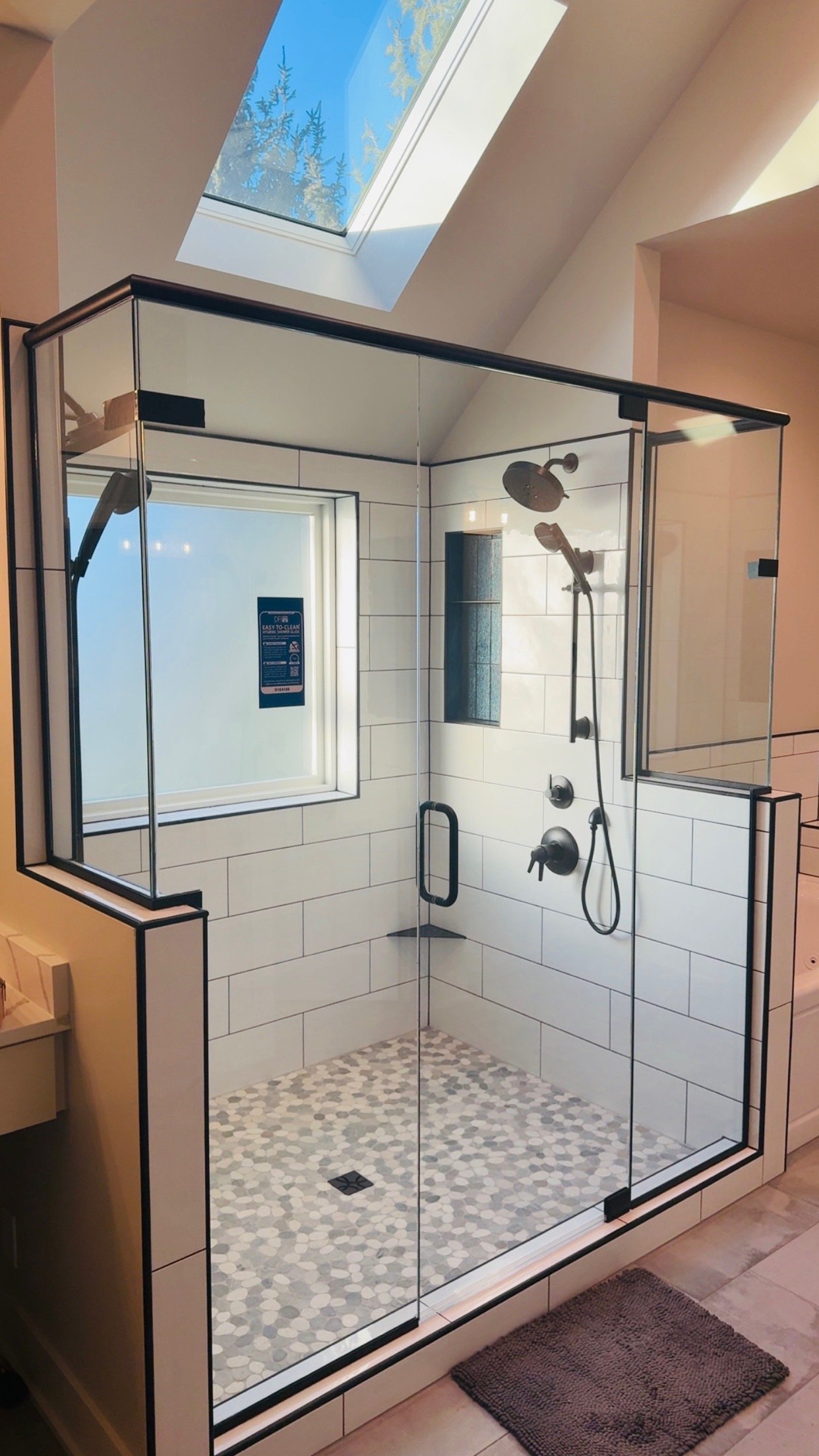 A modern shower enclosure with clear glass walls, black trim, and a pebble stone shower floor. It features a rain shower head and a handheld shower head, with a small window and skylight above.