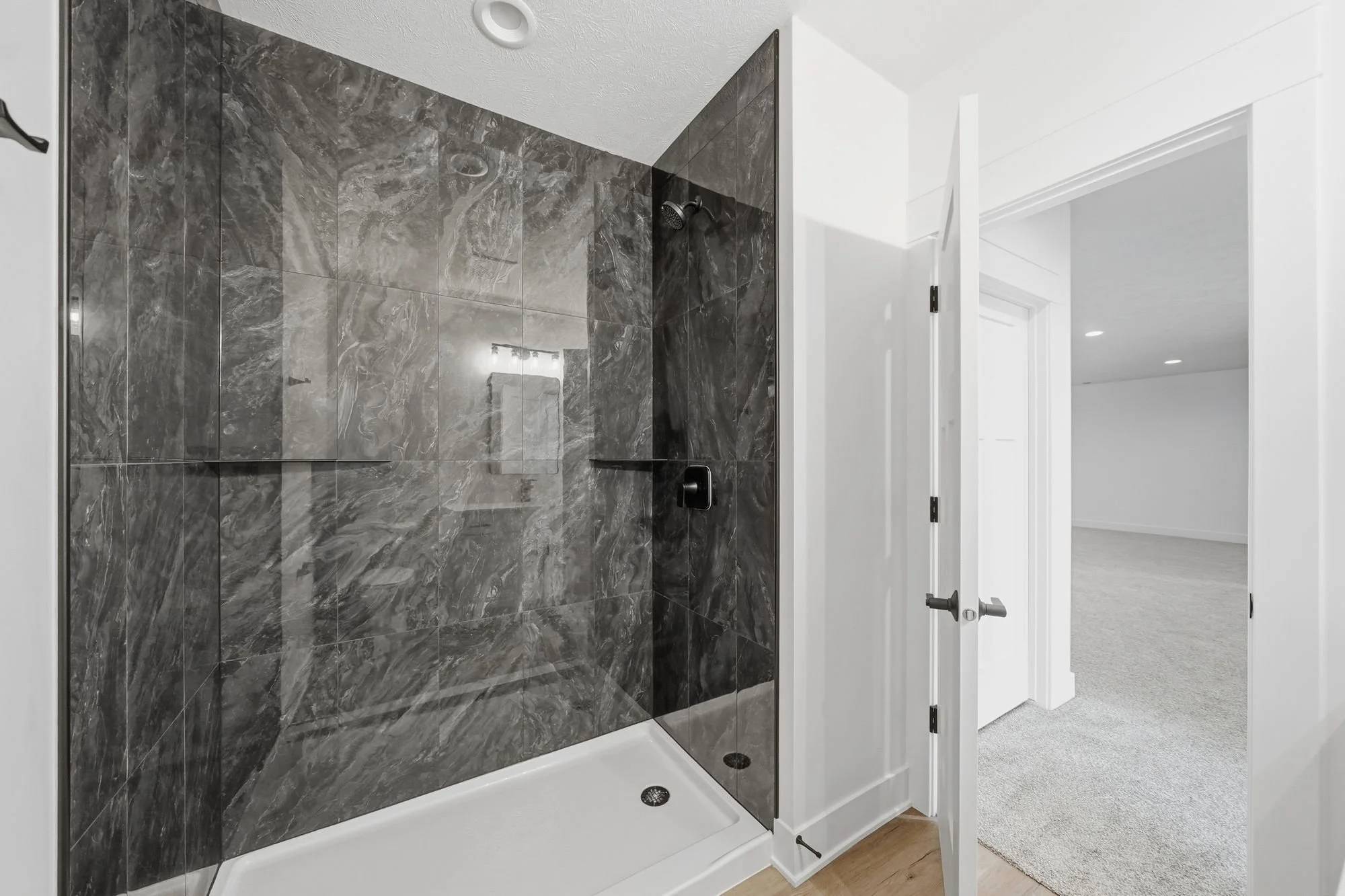 A modern walk-in shower with dark gray marble tiles, a black showerhead, and a glass panel, accessed through a white door in a bright room.