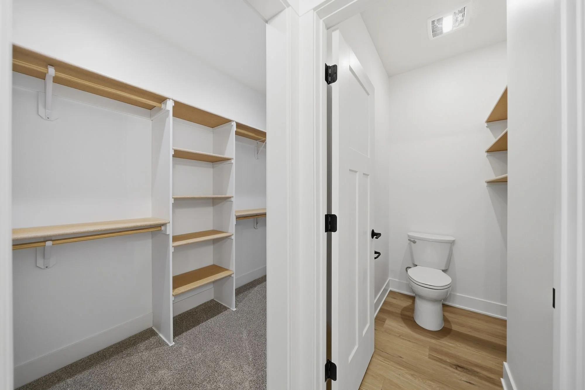 View of a walk-in closet with wooden shelves and hanging space on the left, and a bathroom with a toilet and a vent on the ceiling visible through a partially open door on the right.