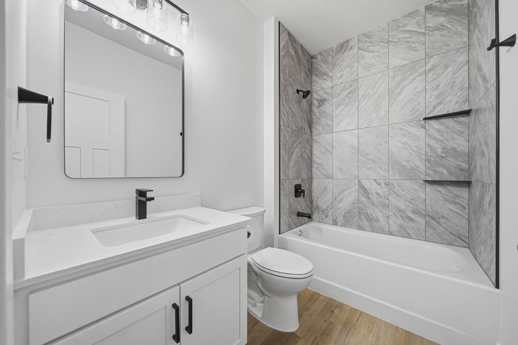 Modern bathroom with white vanity, black fixtures, wall mirror, toilet, and a bathtub with a marble-tiled shower area.