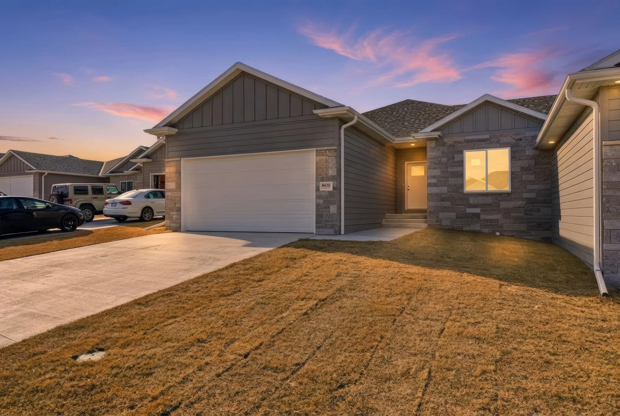 A residential neighborhood at sunset with a modern house featuring gray siding, stone accents, and a white garage door. Several parked cars are visible in the driveway, and the sky is painted with pink and purple hues.