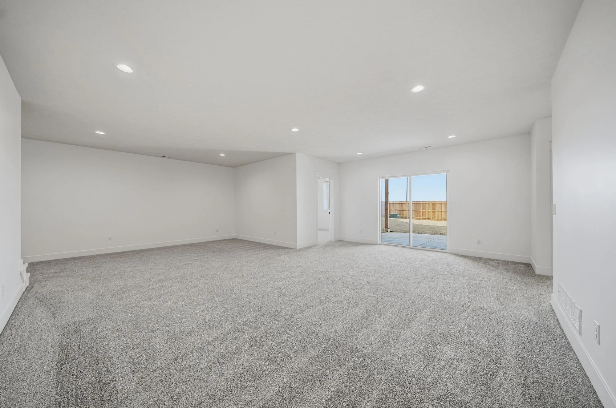 Empty living room with white walls, beige carpet flooring, recessed ceiling lights, sliding glass door leading outdoors, and small windows.