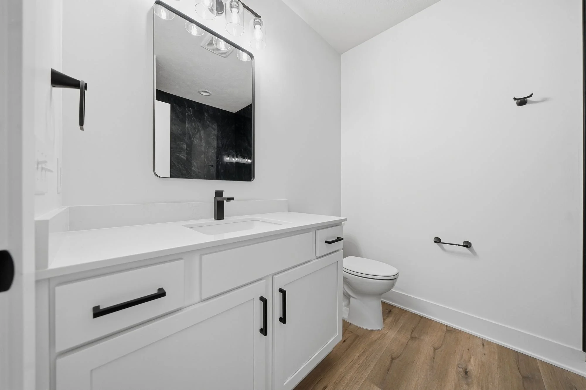 Modern bathroom with white vanity, black fixtures, rectangular mirror, black and clear light fixtures, toilet, and wood flooring.