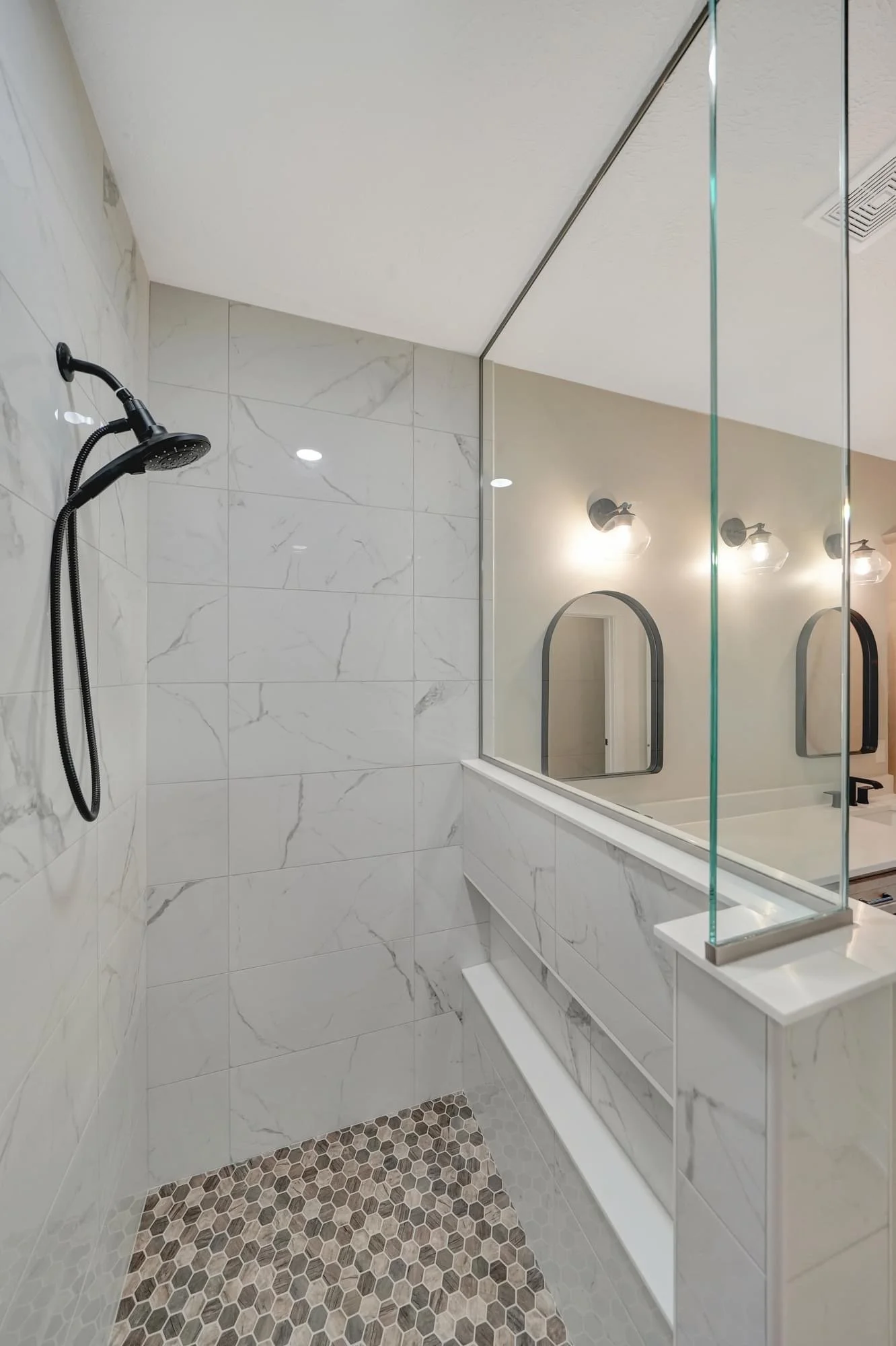 Modern bathroom with white marble tile walls, a black showerhead, a large mirror, and hexagonal floor tiles.