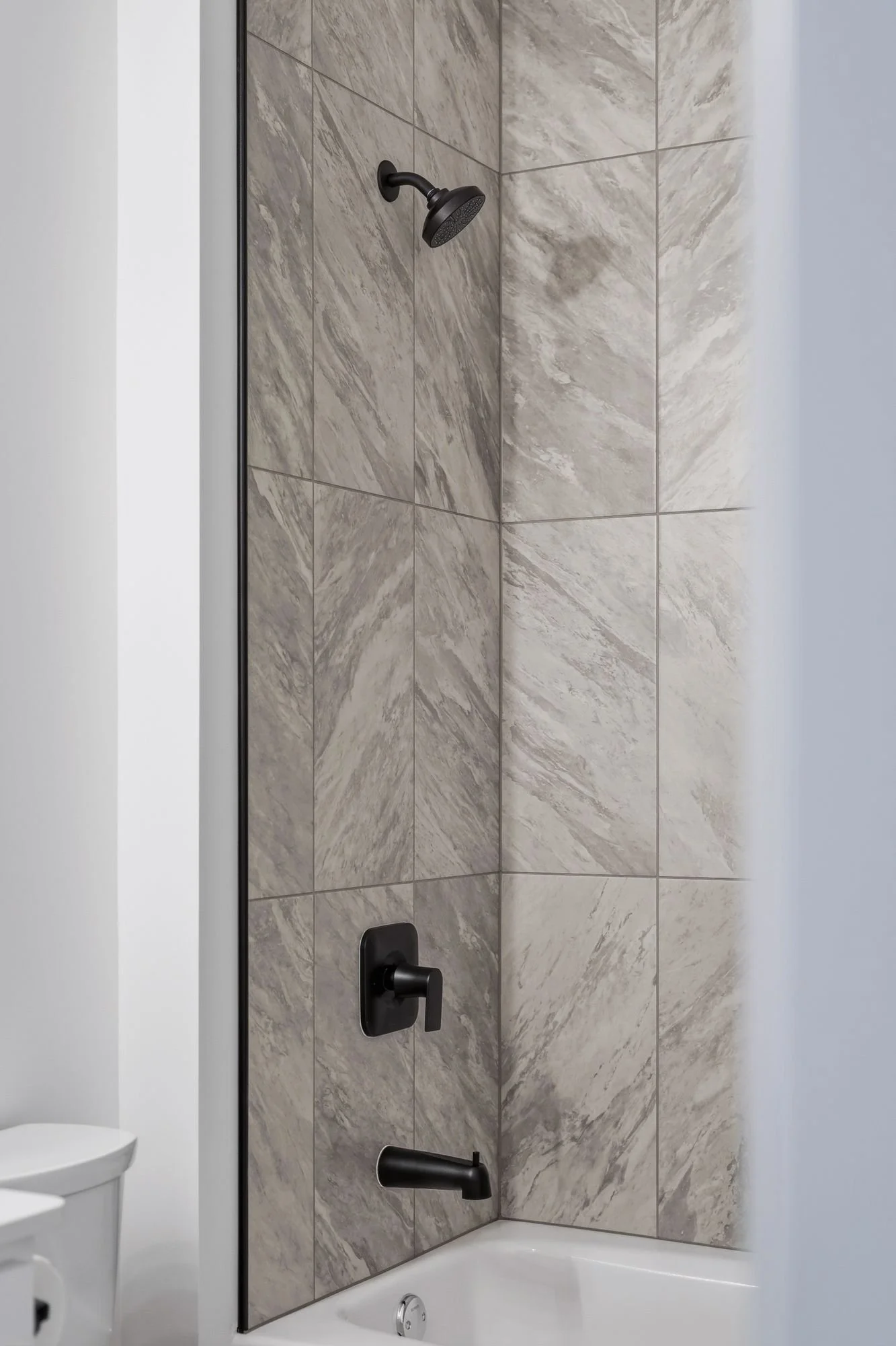 A bathroom with a shower area featuring light gray marble tiles, a black wall-mounted showerhead, black faucet handles, and a white bathtub with a silver drain cover.