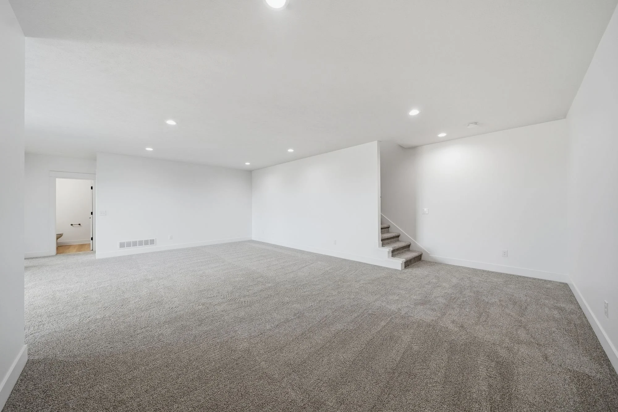 Empty living room with white walls, gray carpet, and staircase with carpeted steps.