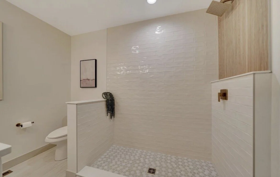 Modern bathroom with a walk-in shower, white brick tiles on the walls, wooden paneling, and a ceiling-mounted showerhead. There is a partial wall separating the shower from a toilet area, which has a framed art piece on the wall and a toilet paper ho