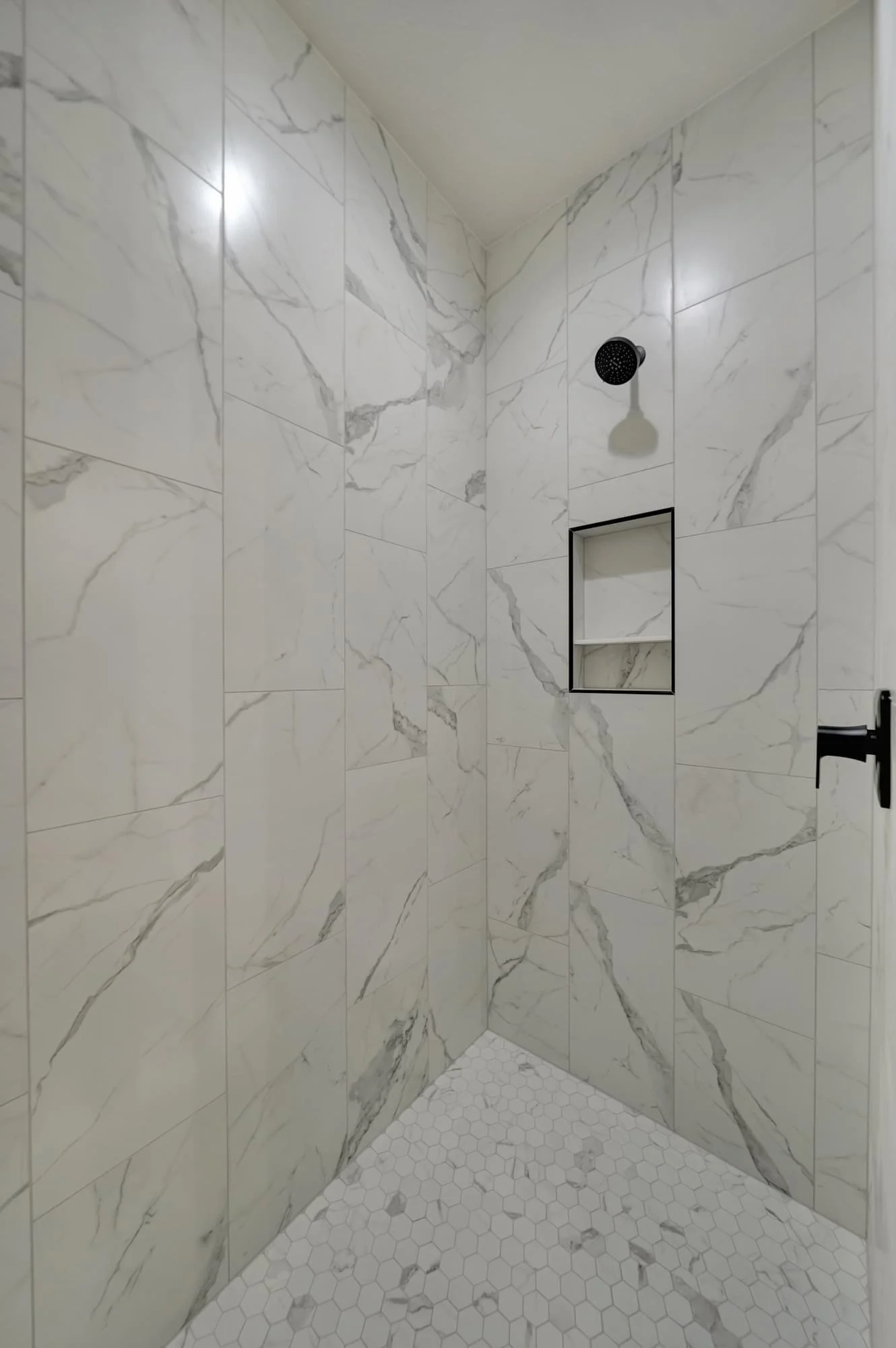 A modern walk-in shower with large white marble tiles, built-in shelves, a black rainfall showerhead, and light hexagonal floor tiles.