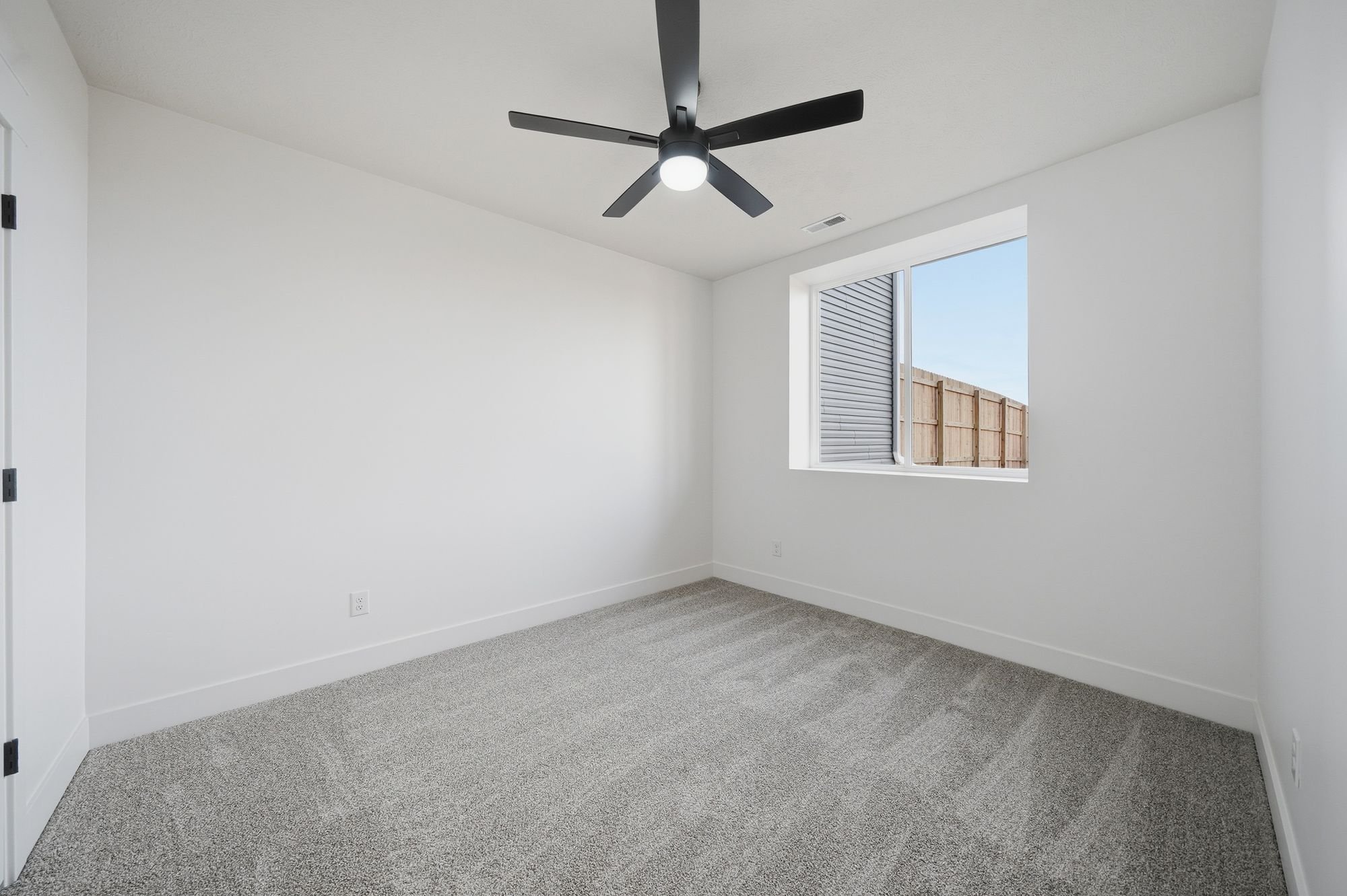 Empty white room with a window showing a wooden fence outside and a black ceiling fan with lights.