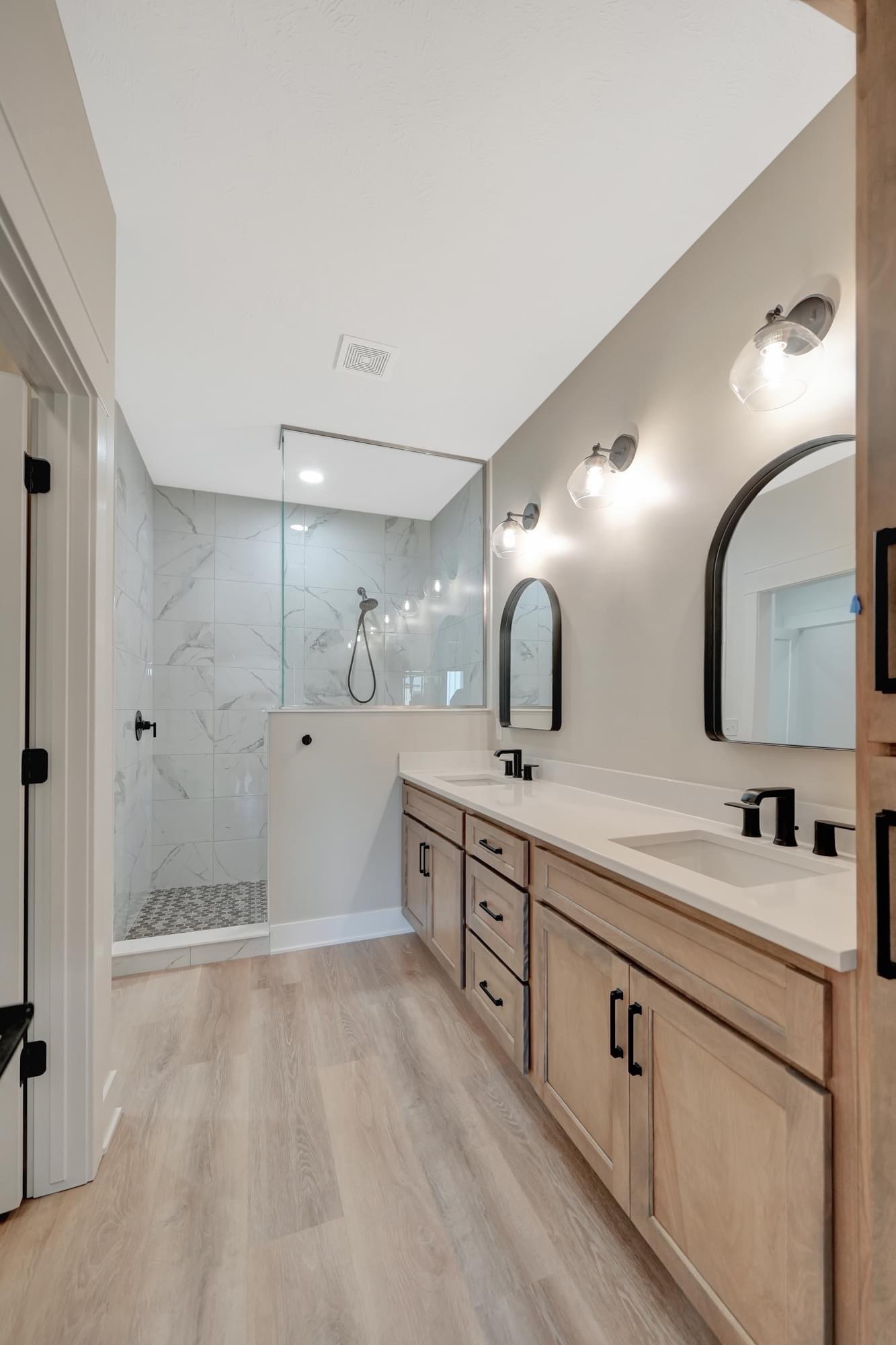 Modern bathroom with dual sinks, wooden cabinets, large mirrors, wall-mounted lights, and a walk-in shower with marble tiles and a glass partition.