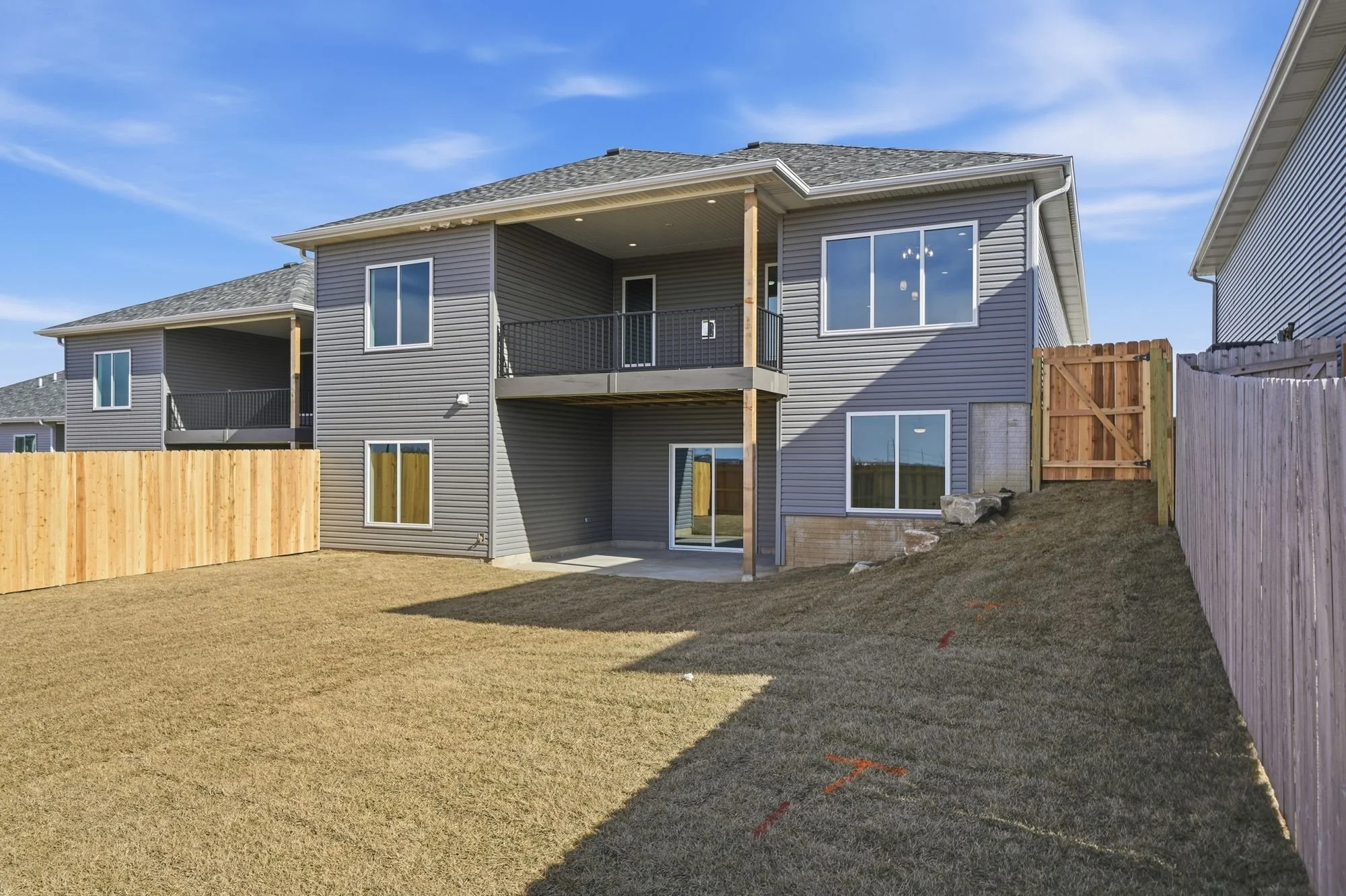 A multi-story house with gray siding, multiple windows, a balcony, and a backyard with a grassy lawn and wooden fences on both sides, under a blue sky.