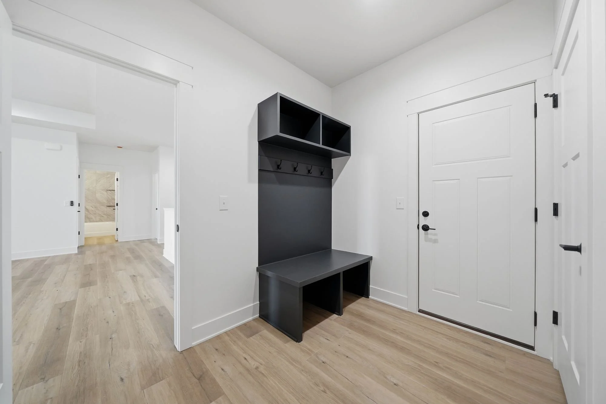 Entryway with white walls, a white door, and a black wall-mounted storage unit with hooks, hardwood flooring, and a view into other rooms.