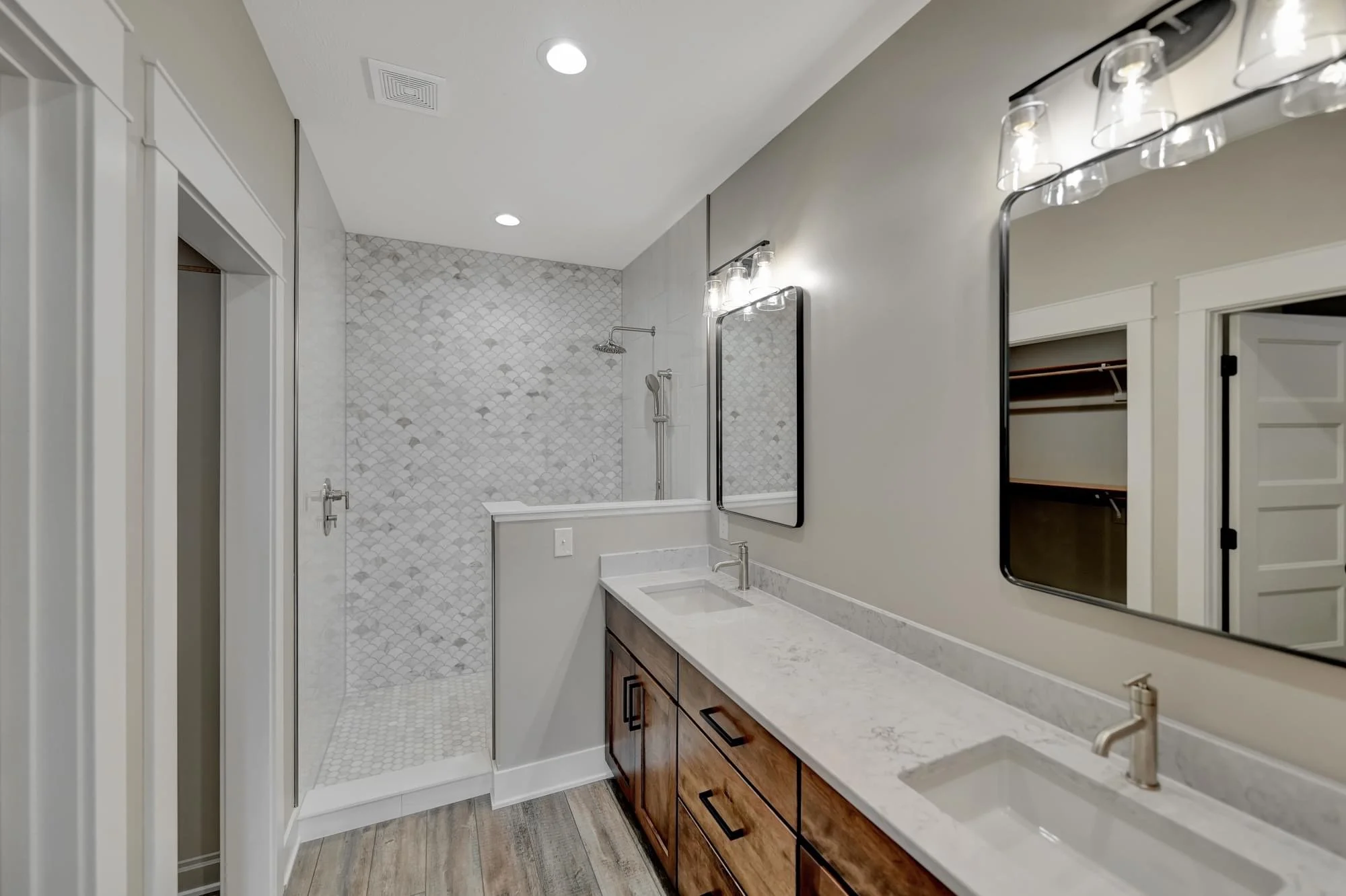 Modern bathroom with a wooden vanity, white marble countertop, large mirror, gray walls, and a walk-in shower with white pebble tile. Two wall-mounted light fixtures with glass shades are above the mirror, and recessed ceiling lights are present.