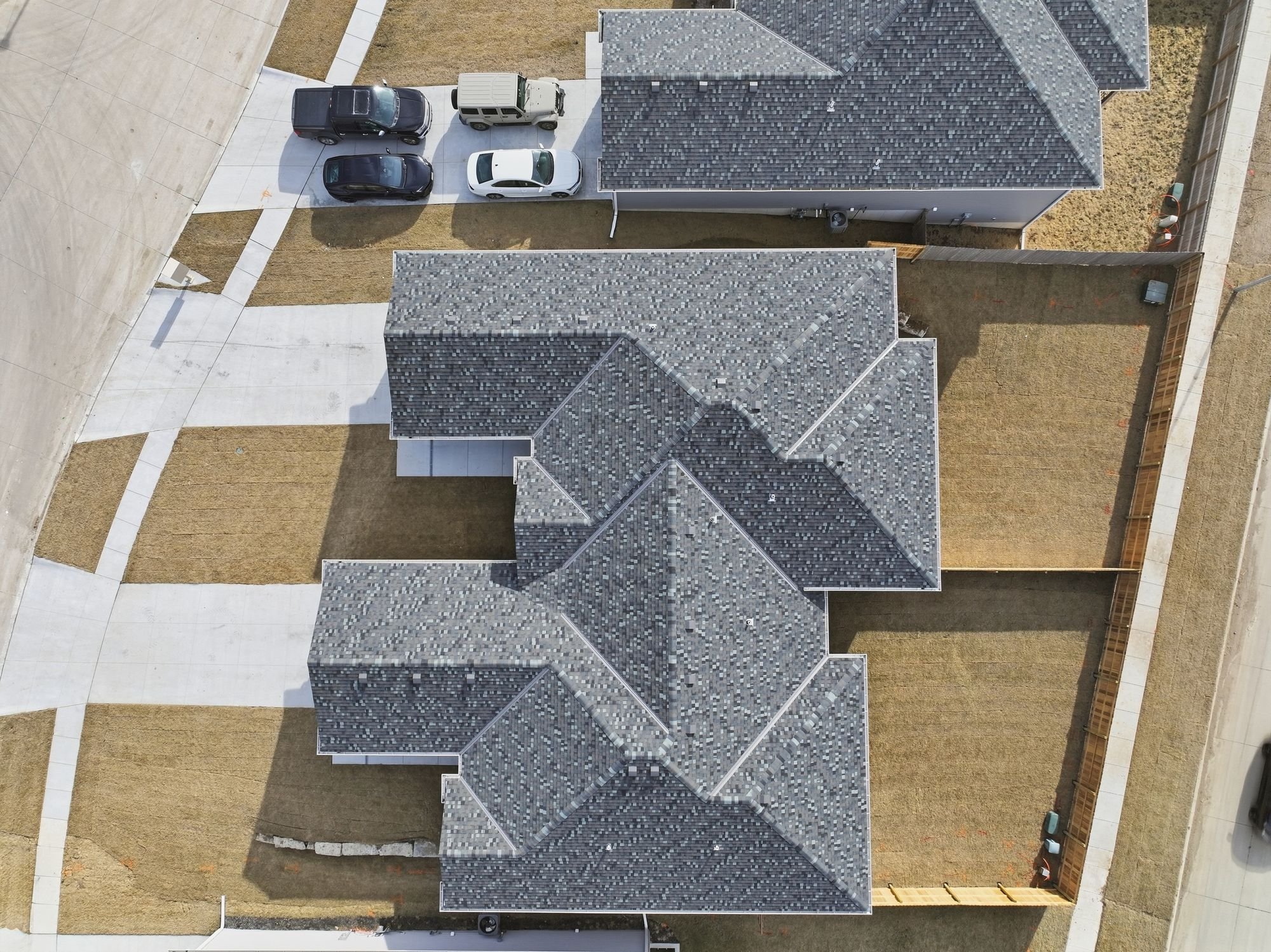 An aerial view of a modern residential house with a gray shingle roof, surrounded by a small yard with brown grass, a concrete driveway, and three parked cars.