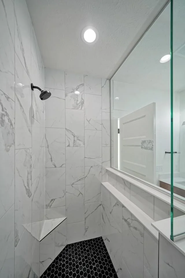 Modern bathroom shower with white marble walls, black hexagonal tile floor, and a large mirror reflecting the ceiling lights.