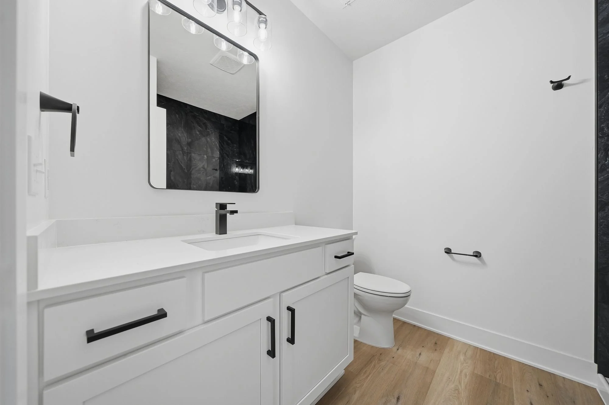 Modern bathroom with white vanity, black hardware, black-framed mirror, black light fixture, black towel hook, toilet, wood flooring, and white walls.