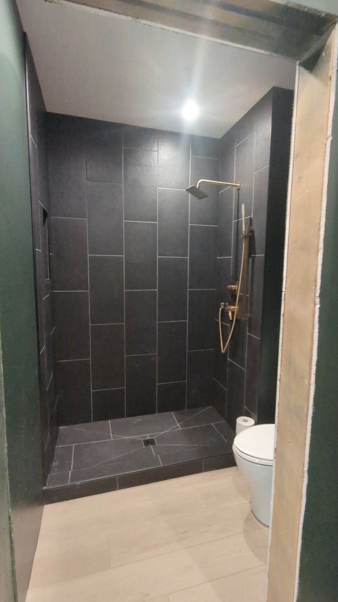 A modern shower area with black tiles on the walls and floor, a rainfall showerhead, and a handheld shower with a control panel, located in a bathroom with a toilet and a roll of toilet paper.